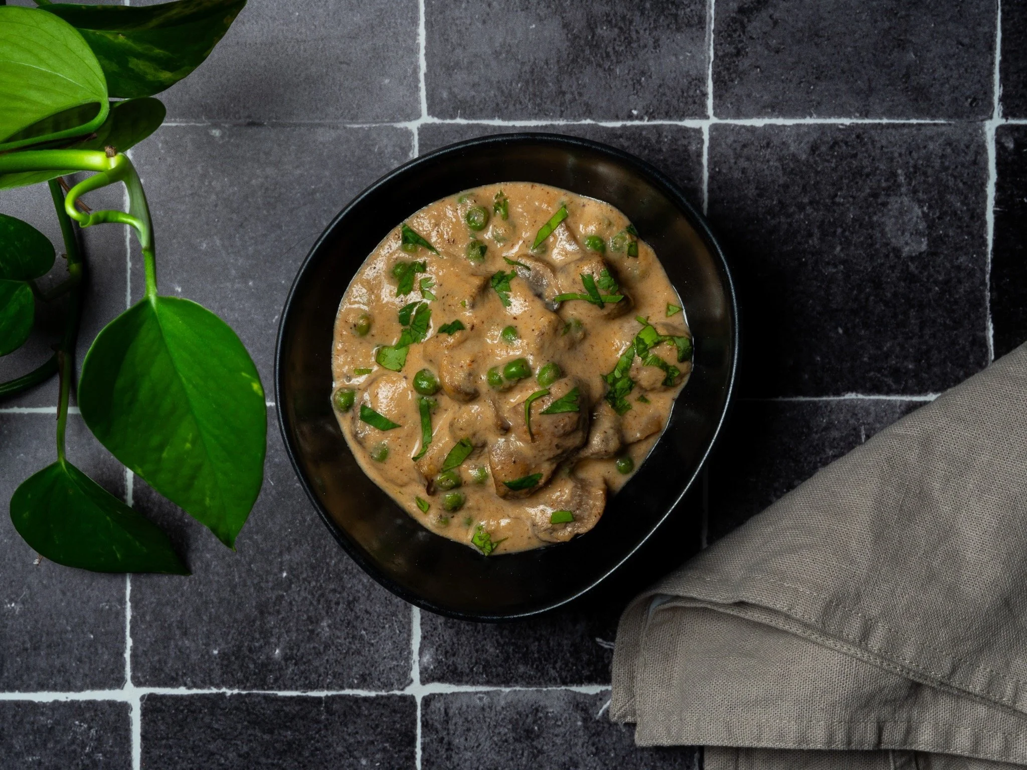 Guiso de carne con salsa cremosa y guisantes en un plato negro, al lado de una planta de hojas verdes y un paño beige, sobre azulejos oscuros.