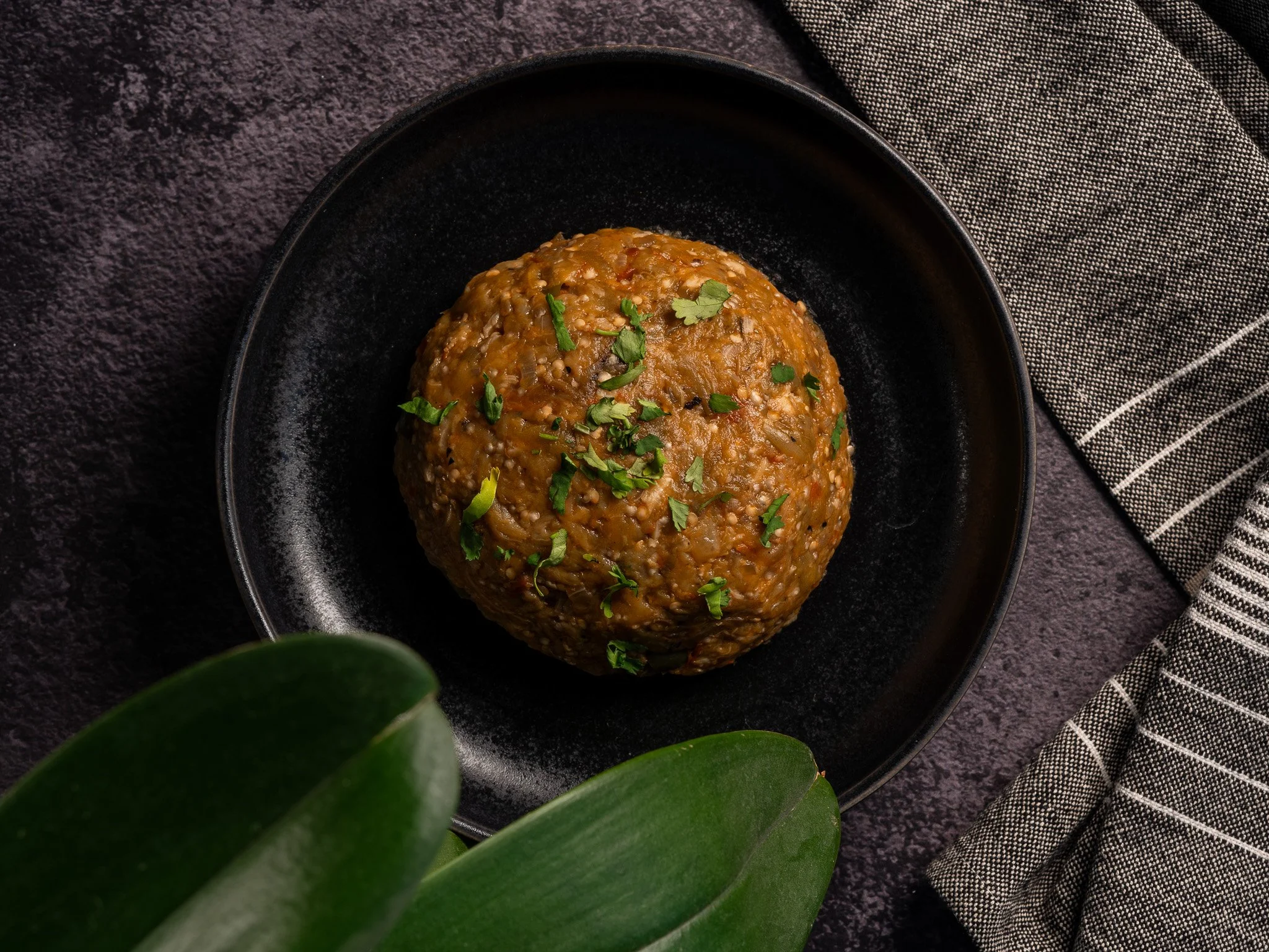 Albóndiga de carne con verduras y arroz en un plato negro, decorada con cilantro, en un fondo oscuro con un paño de tela a rayas y hojas verdes en primer plano.
