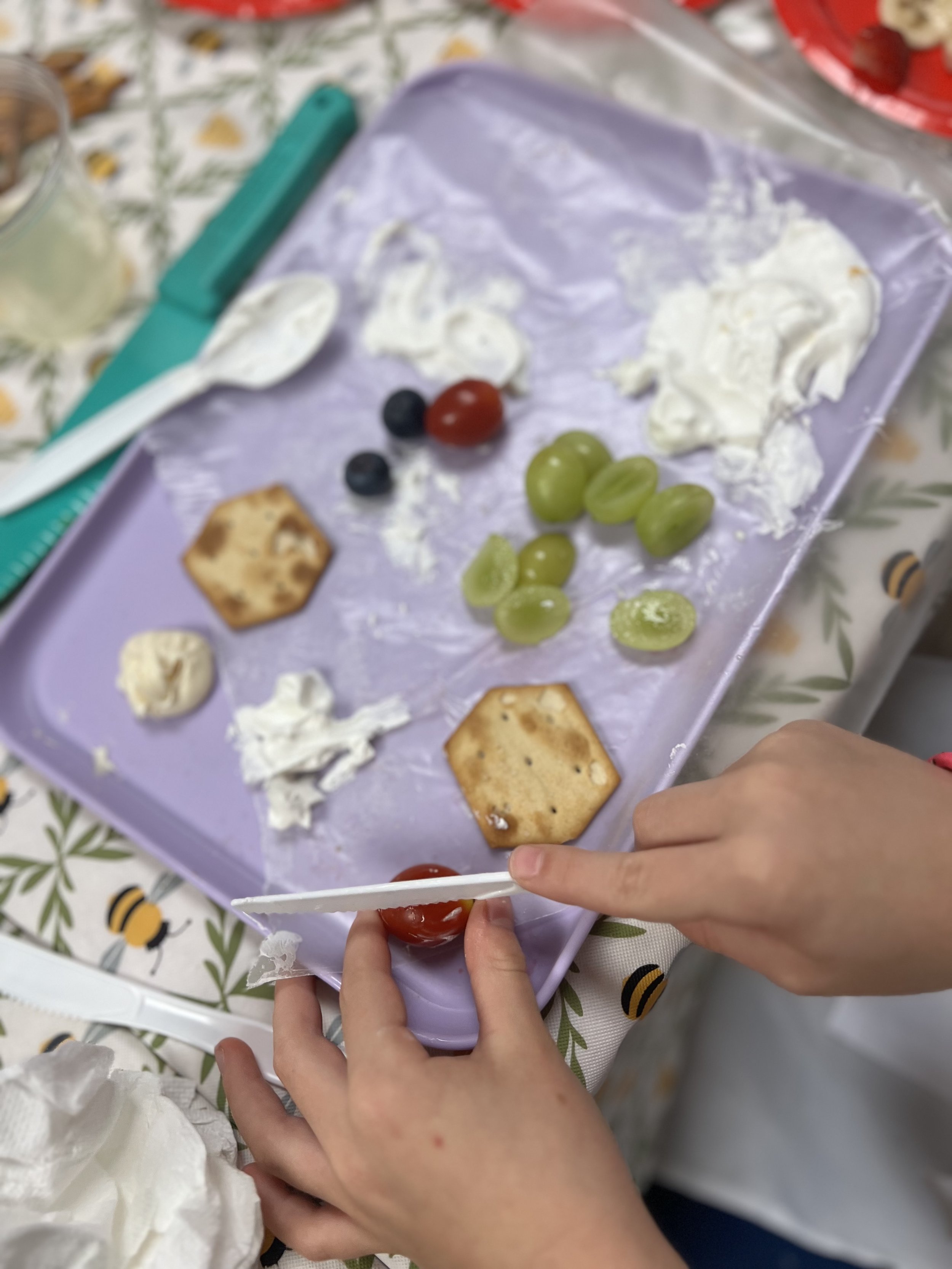 A child's hands holding a plastic knife and a tomato on a tray with whipped cream, crackers, grapes, blueberries, and cherry tomatoes.