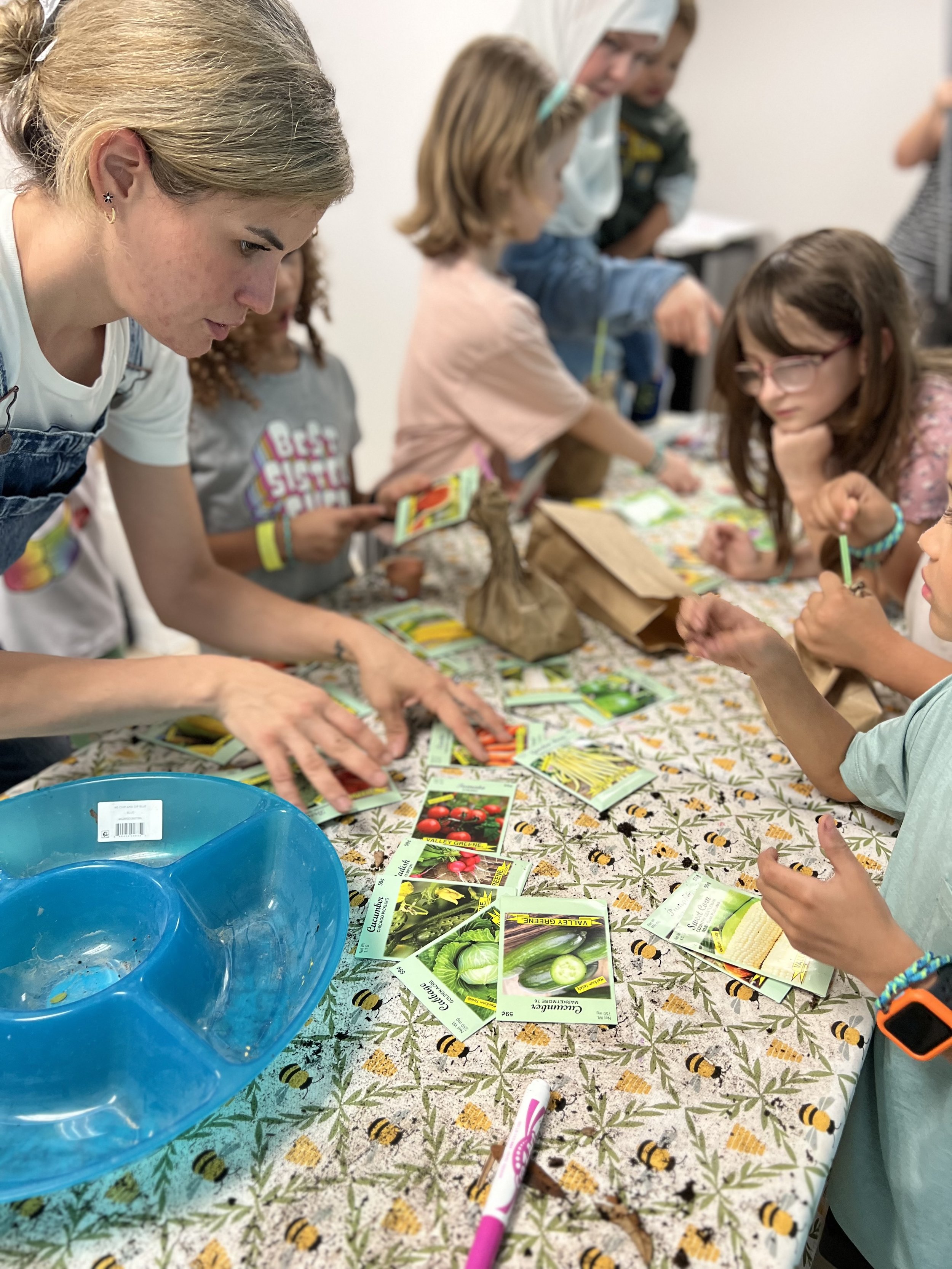 Children and adults sorting seed packets at a table with a bee-patterned tablecloth, engaging in a gardening activity indoors.