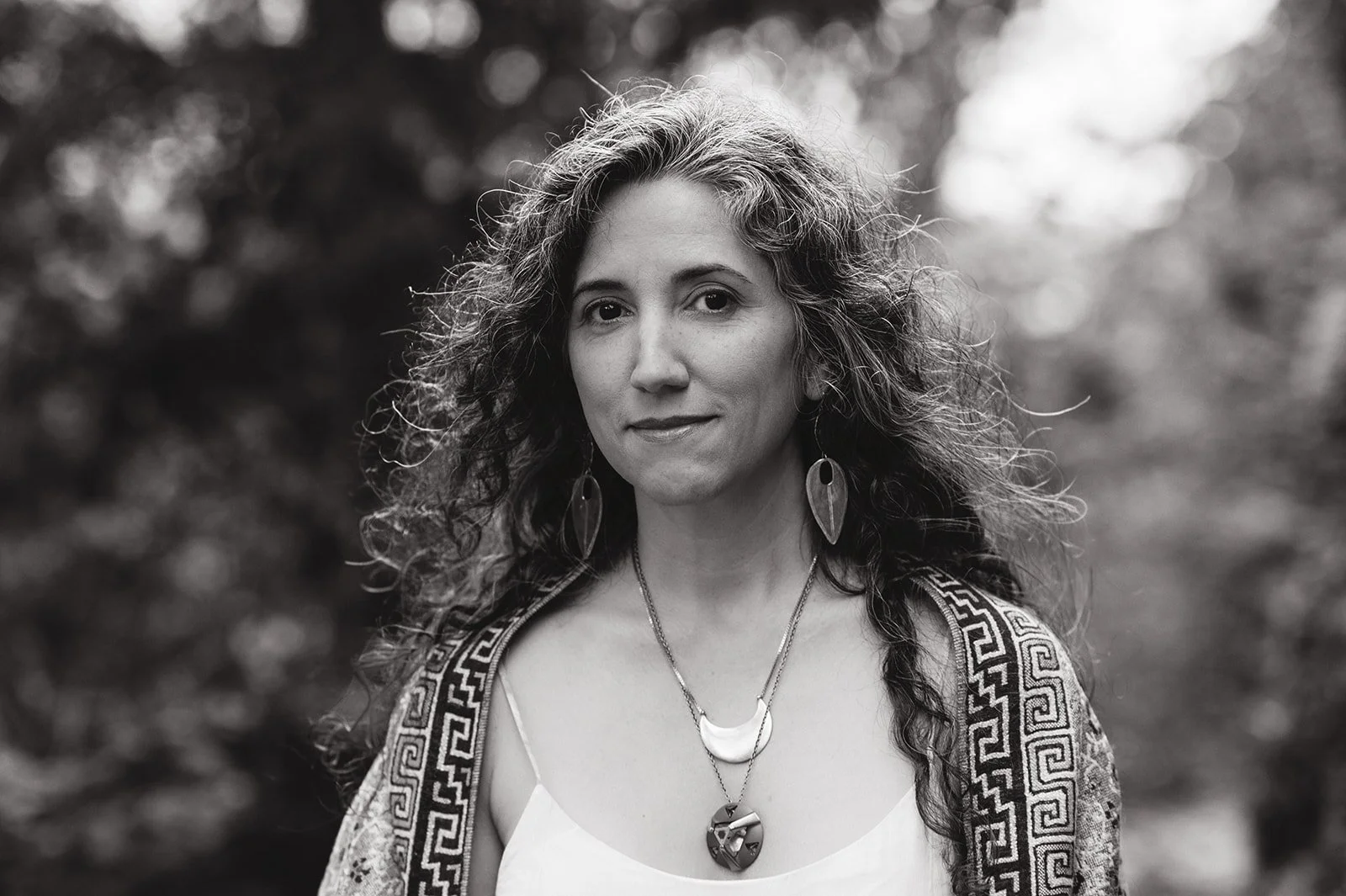 Black and white portrait of a woman with curly hair, wearing earrings and layered necklaces, looking at the camera with a neutral expression, outdoors with blurred background.