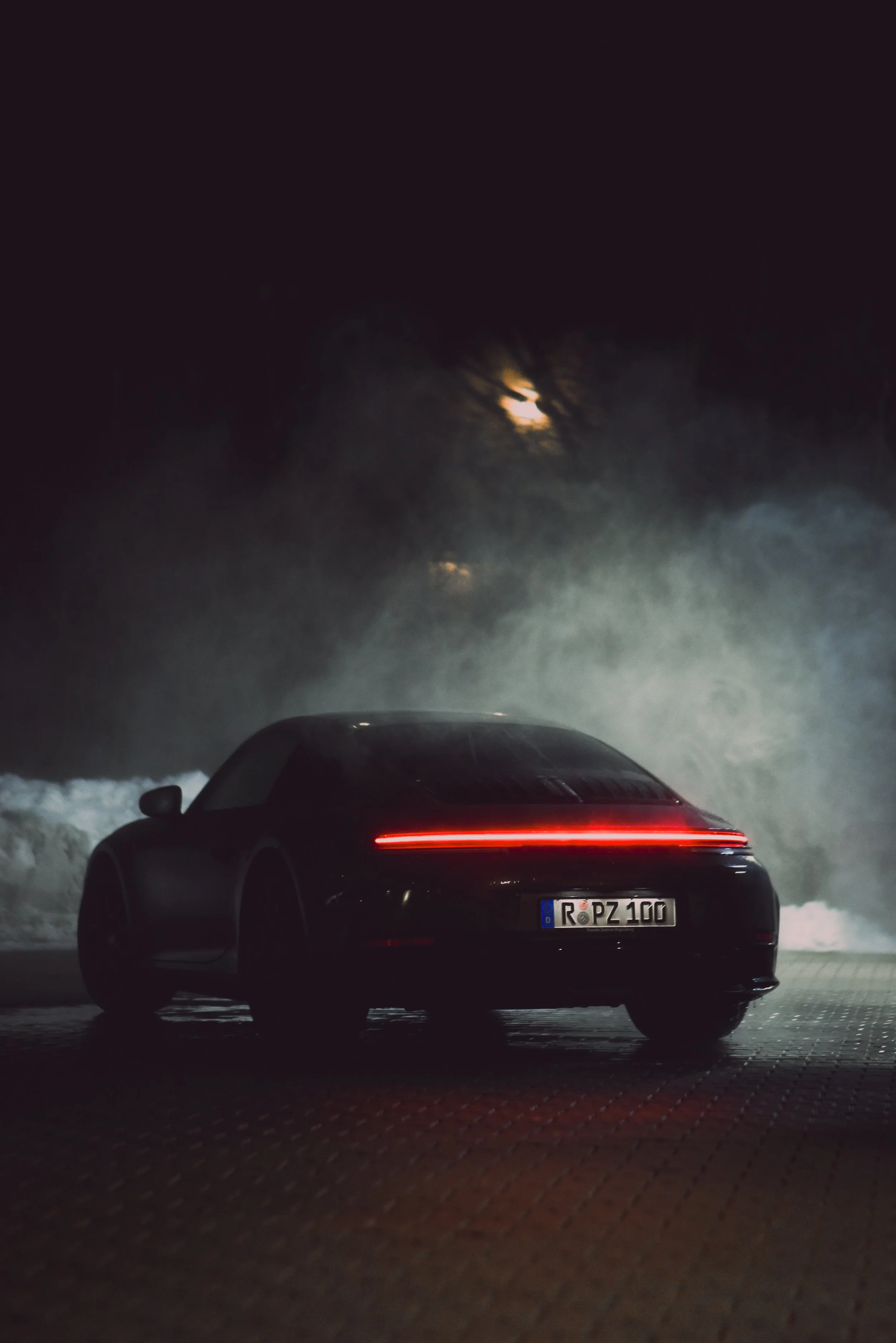 Rear view of a black Porsche 911 sports car at night, illuminated by its rear lights, with mist and waves in the background under a moonlit sky.