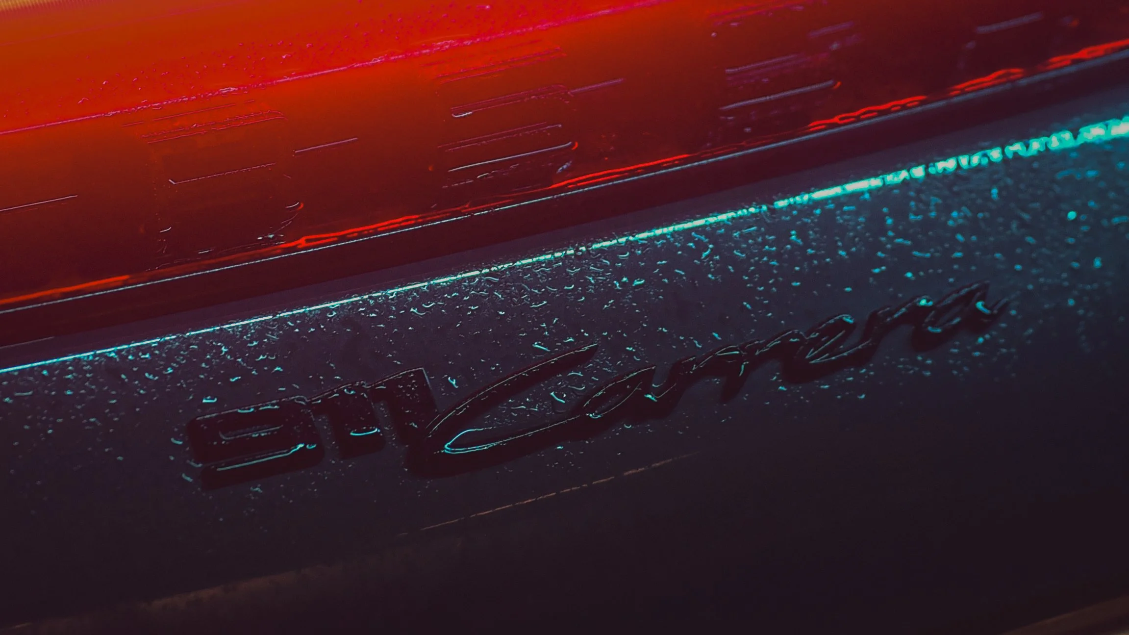 Close-up of a Porsche 911 Carrera logo on the rear of a car with neon lighting and water droplets.