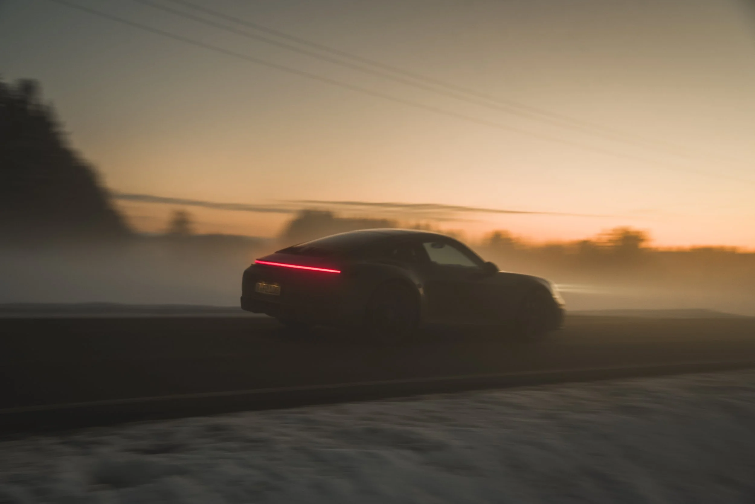 A Porsche 911 Carrera driving on a road at sunset with a blurred background, and a red lighted strip on the rear of the car. sunset