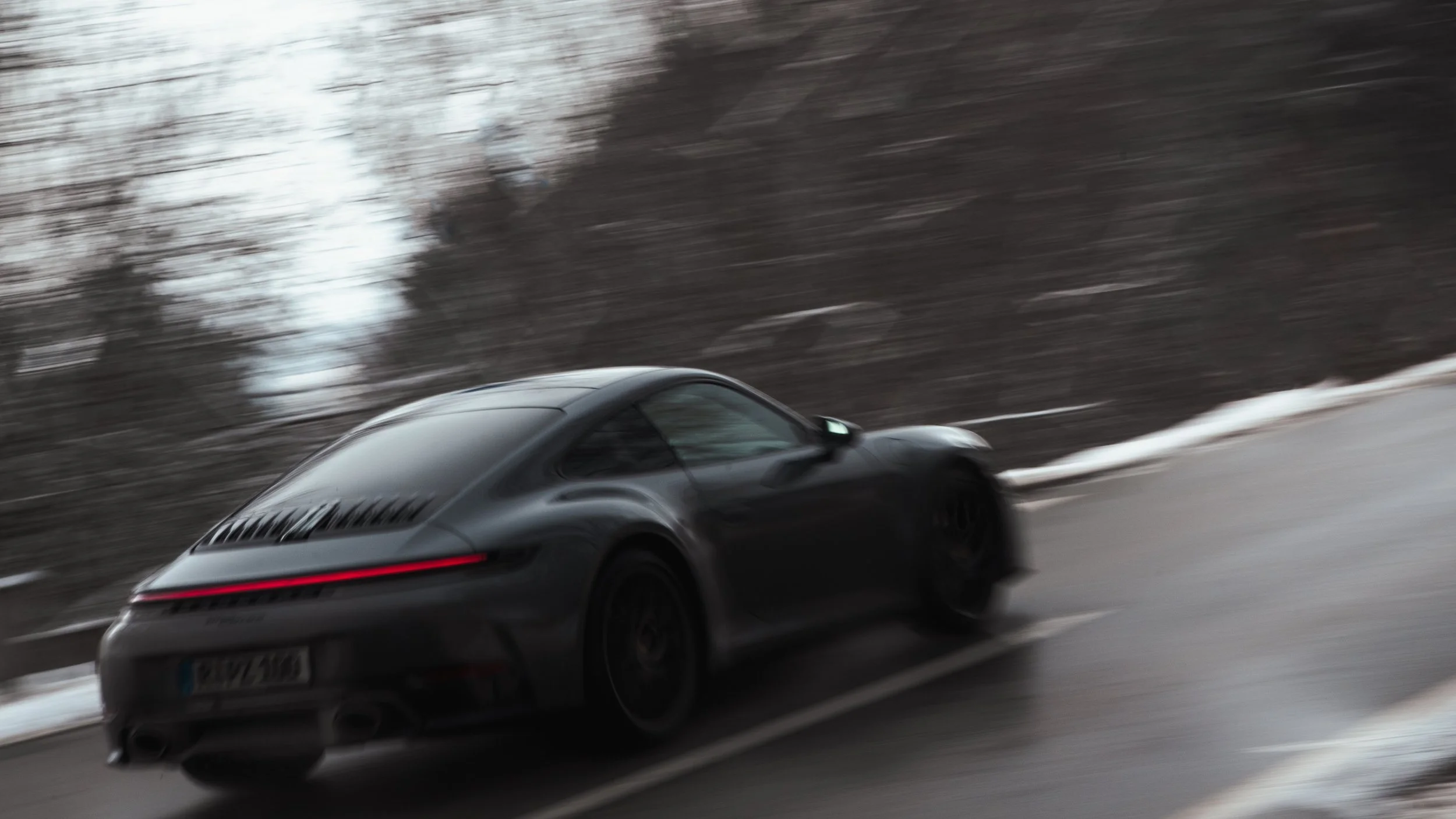 Blurred image of a black Porsche Carrera 911 driving on a wet road with snow  in the background.