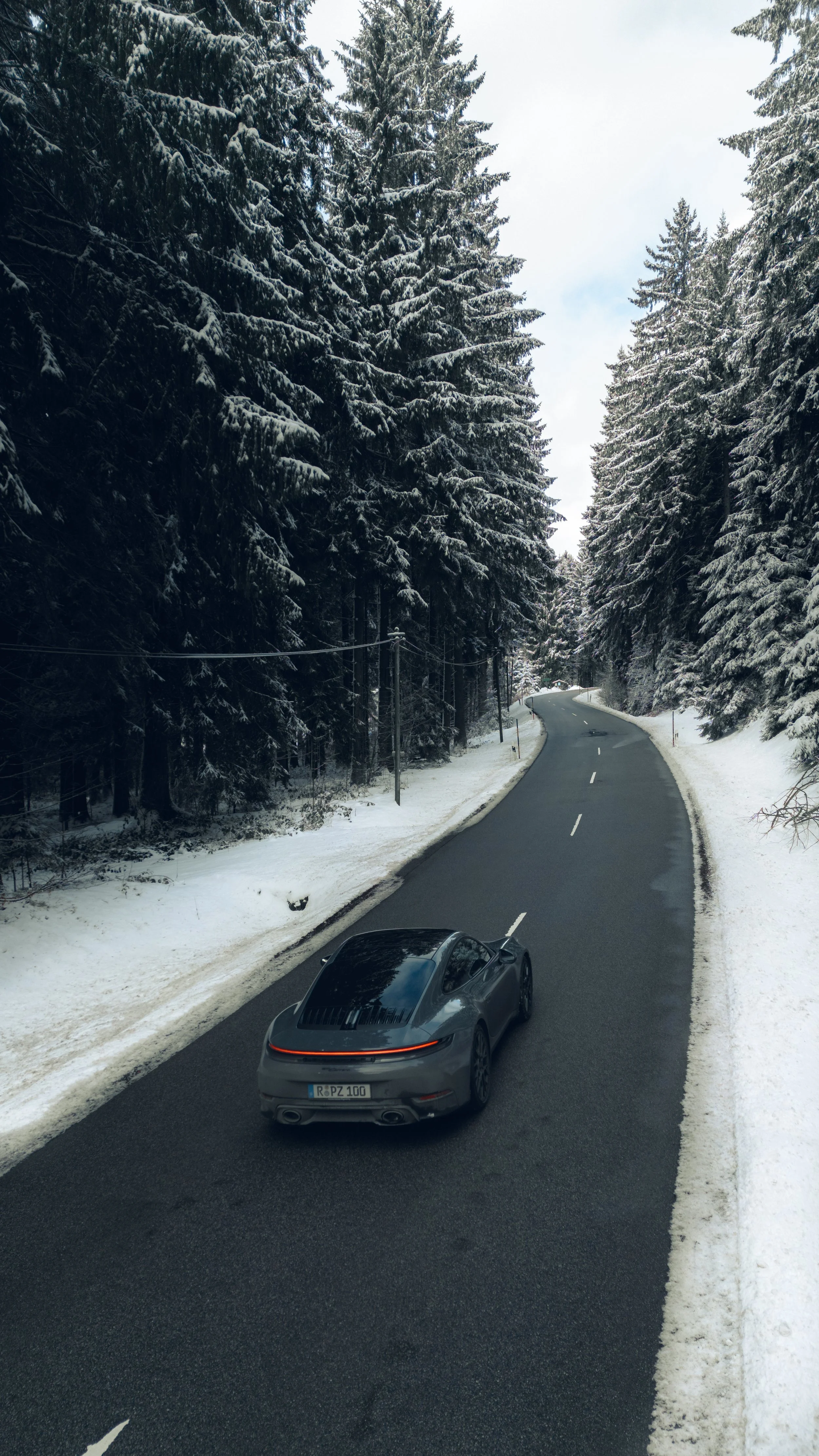 A grey Porsche 911 Carrera driving on a winding mountain road surrounded by snow-covered trees on both sides.