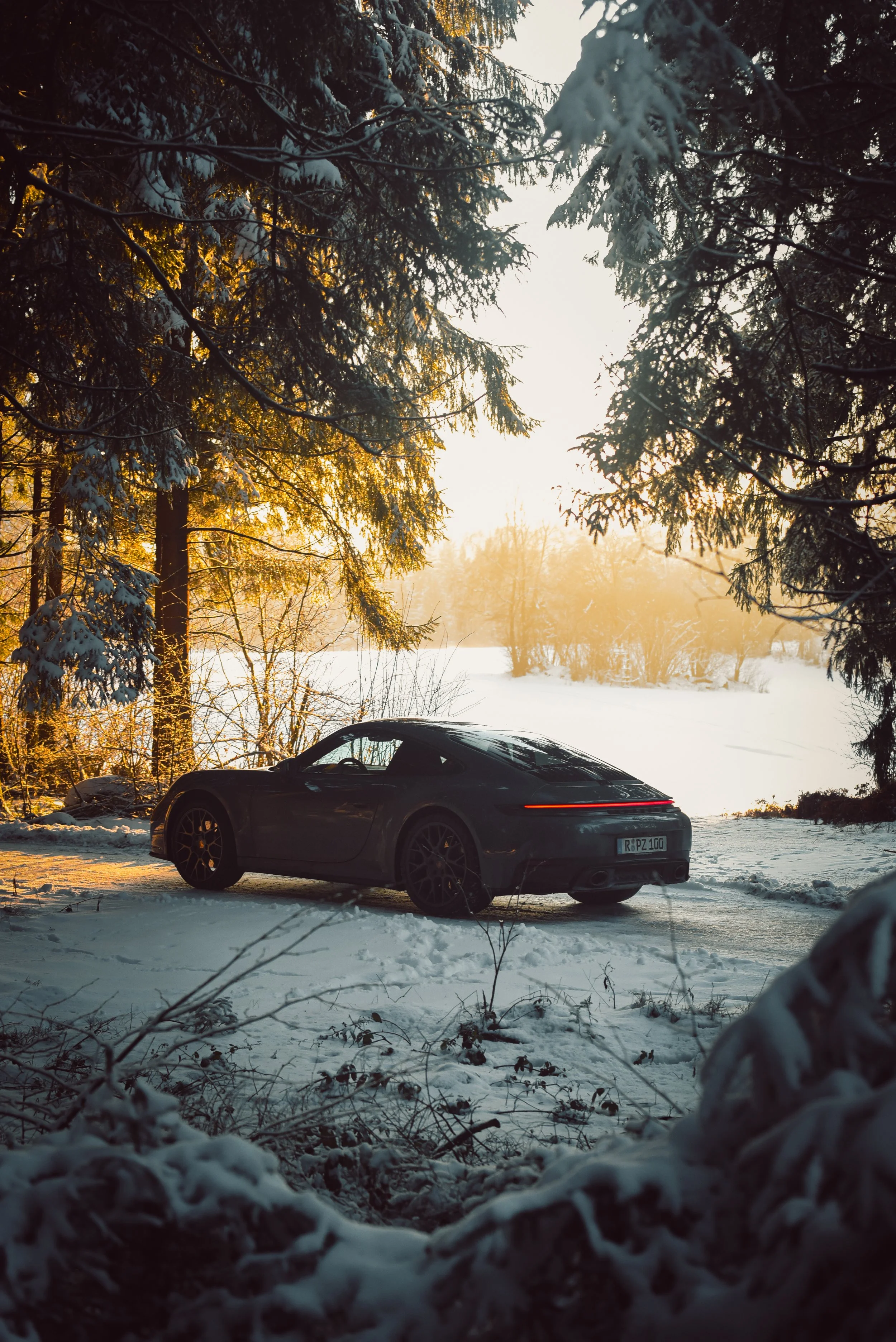 A grey Porsche 911 Carrera car parked on a snowy road during sunset, surrounded by snow-covered trees in a winter landscape.