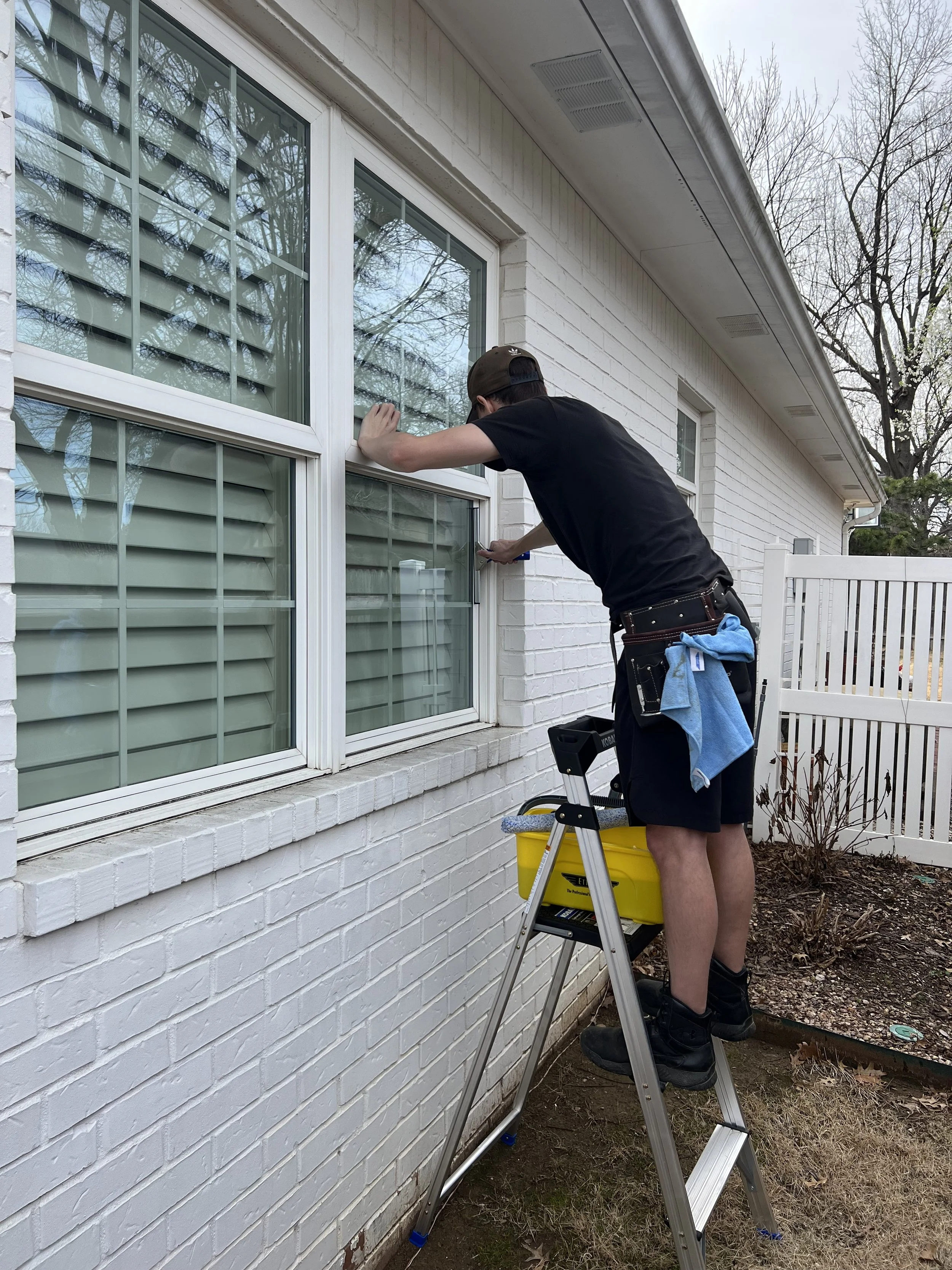 A person using an extension pole to clean or remove debris from the roof of a two-story white house with vinyl siding and a bay window. The scene is outdoors on a sunny day with a blue sky and some clouds.
