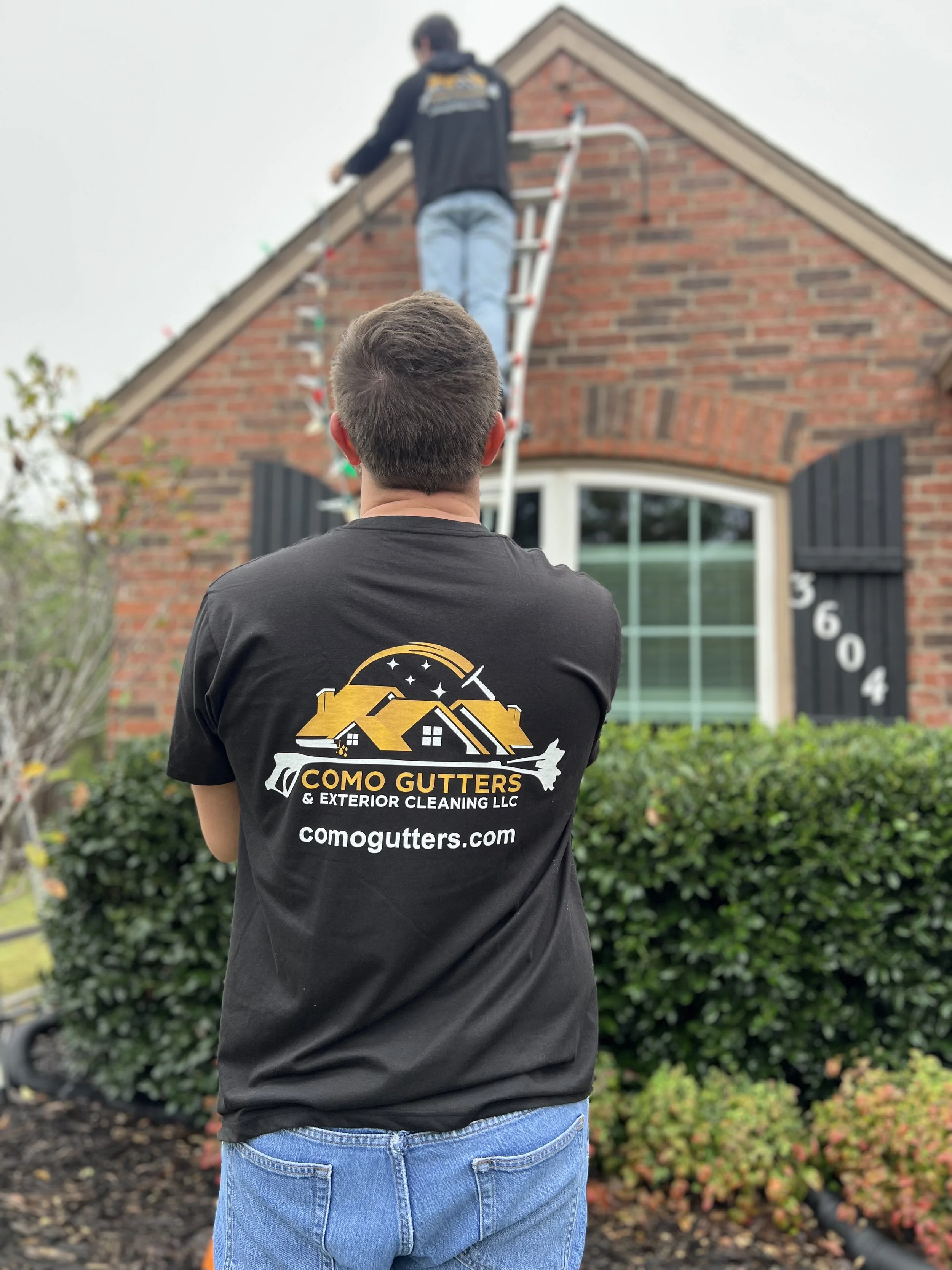 A man with short hair wearing a black Como Gutters company T-shirt standing outside in front of bushes, watching another worker on a ladder cleaning gutters on a brick house.