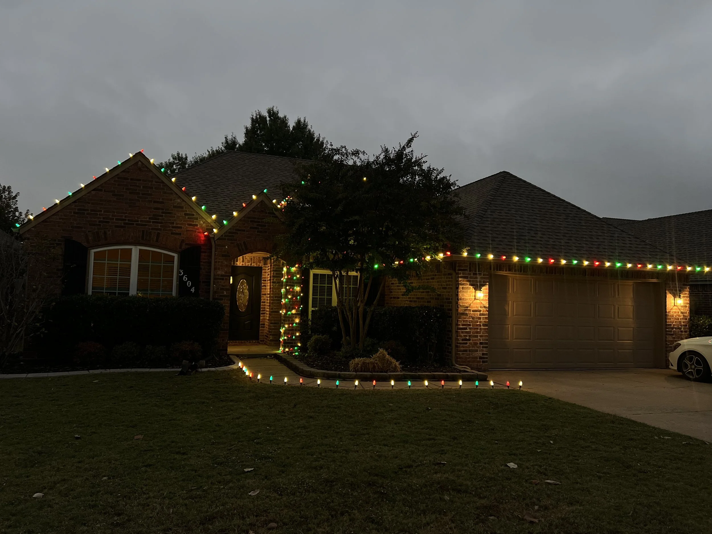 A single-story brick house decorated with multicolored Christmas lights along the roofline, around the front door, and sidewalk. The house is illuminated by wall-mounted exterior lights, with a well-maintained lawn and landscaped bushes in front. The sky is overcast and it is evening.