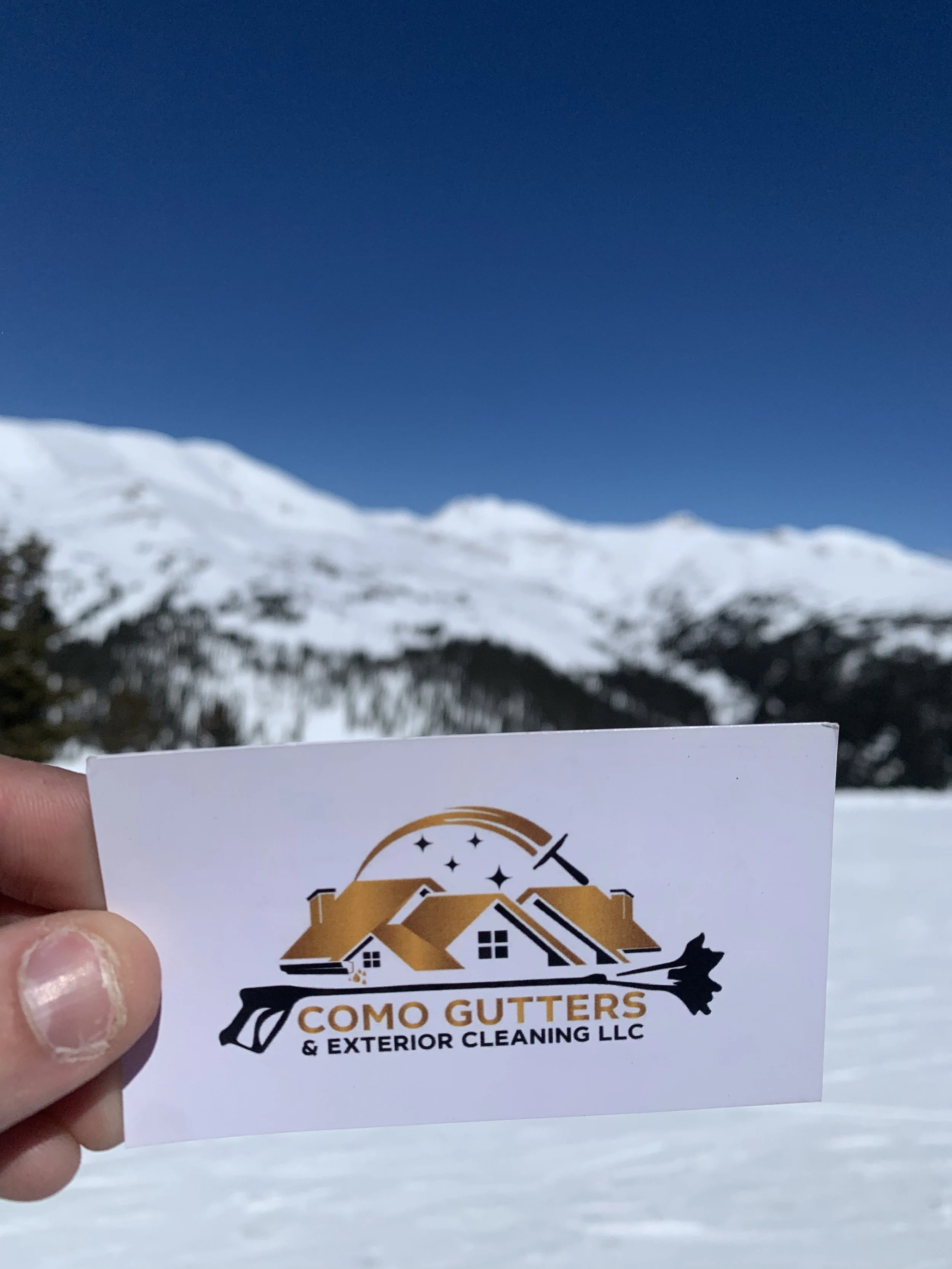 Hand holding a business card for Como Gutters & Exterior Cleaning LLC in front of snowy mountains under a blue sky.