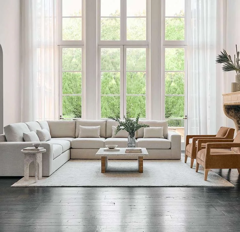 Bright living room with large windows, white sectional sofa, glass coffee table with books and vase, side table, two orange armchairs, and decorative plants.
