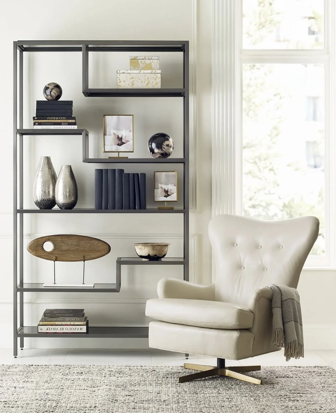 Living room with a tall metal bookshelf filled with decorative objects, books, and framed photos; white armchair with a gray throw blanket, beige carpet, and large window with sunlight.