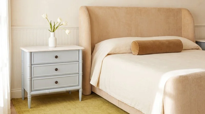 Warm neutral bedroom featuring a tan upholstered bed, cream bedding, a pale grey three-drawer nightstand and white tulips.