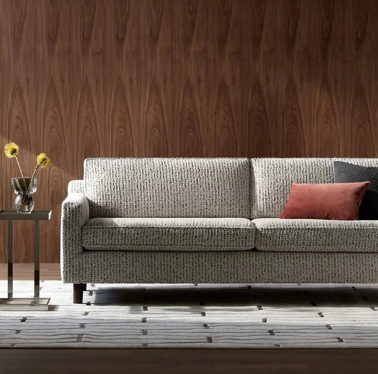 Living room with a beige textured sofa, red and black pillows, a wooden wall, a side table with a vase of yellow flowers, and a striped rug.