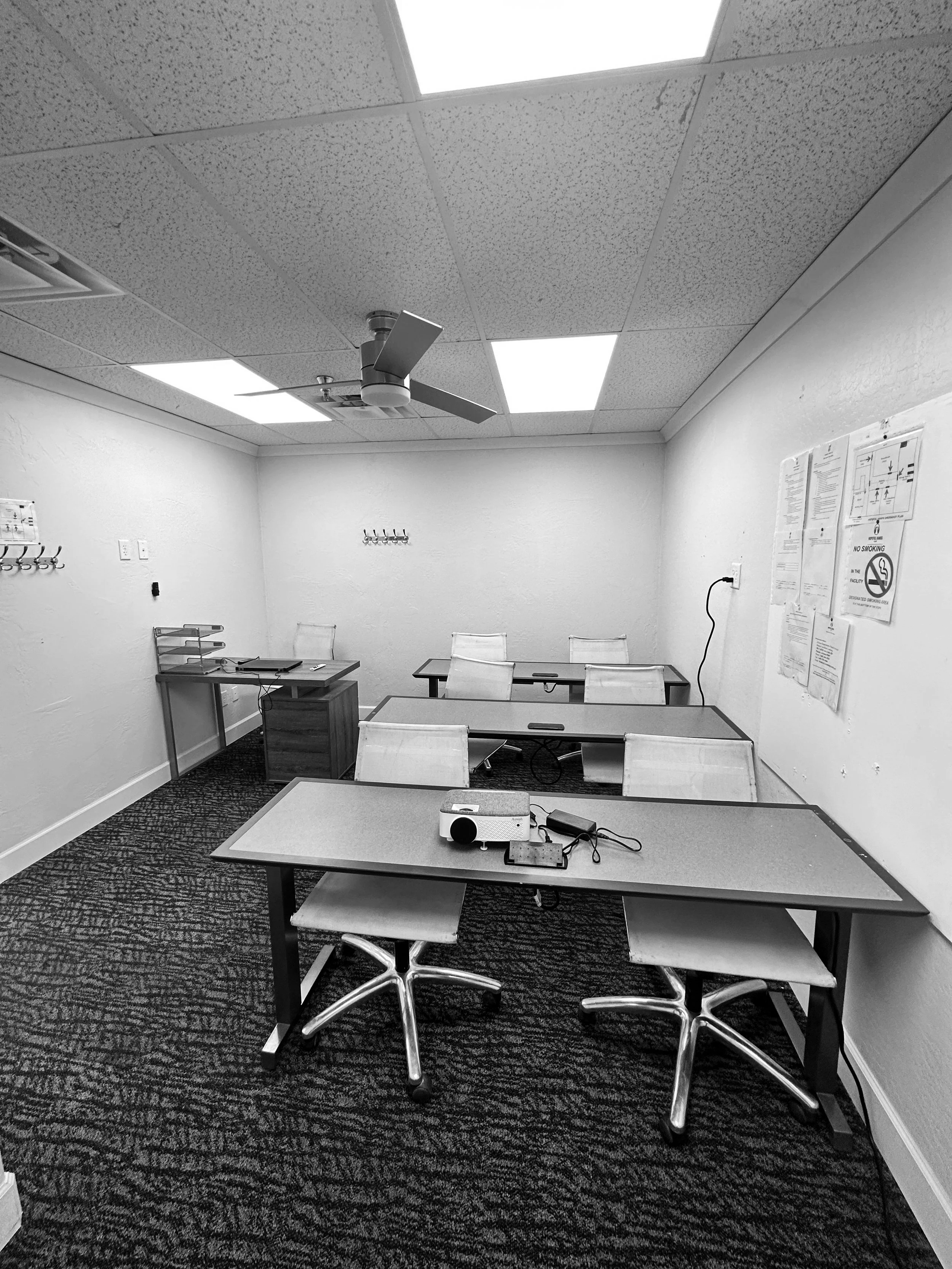 Empty classroom with six beige chairs and three tables, projector on middle table, wall-mounted hooks, posters on wall, and ceiling fan.