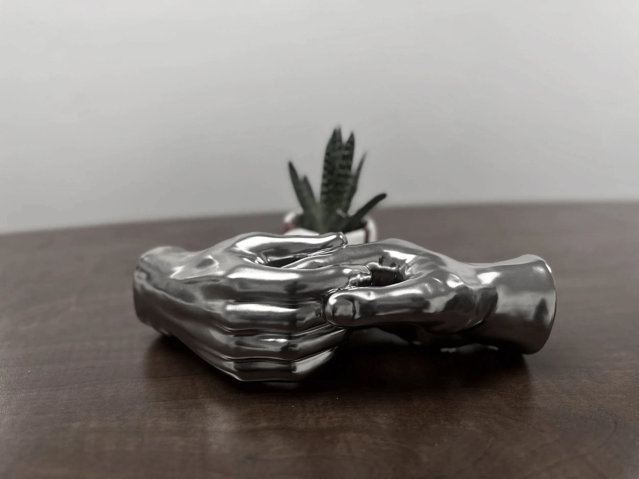 A shiny metallic hand sculpture resting on a wooden surface with a small potted succulent plant in the background.