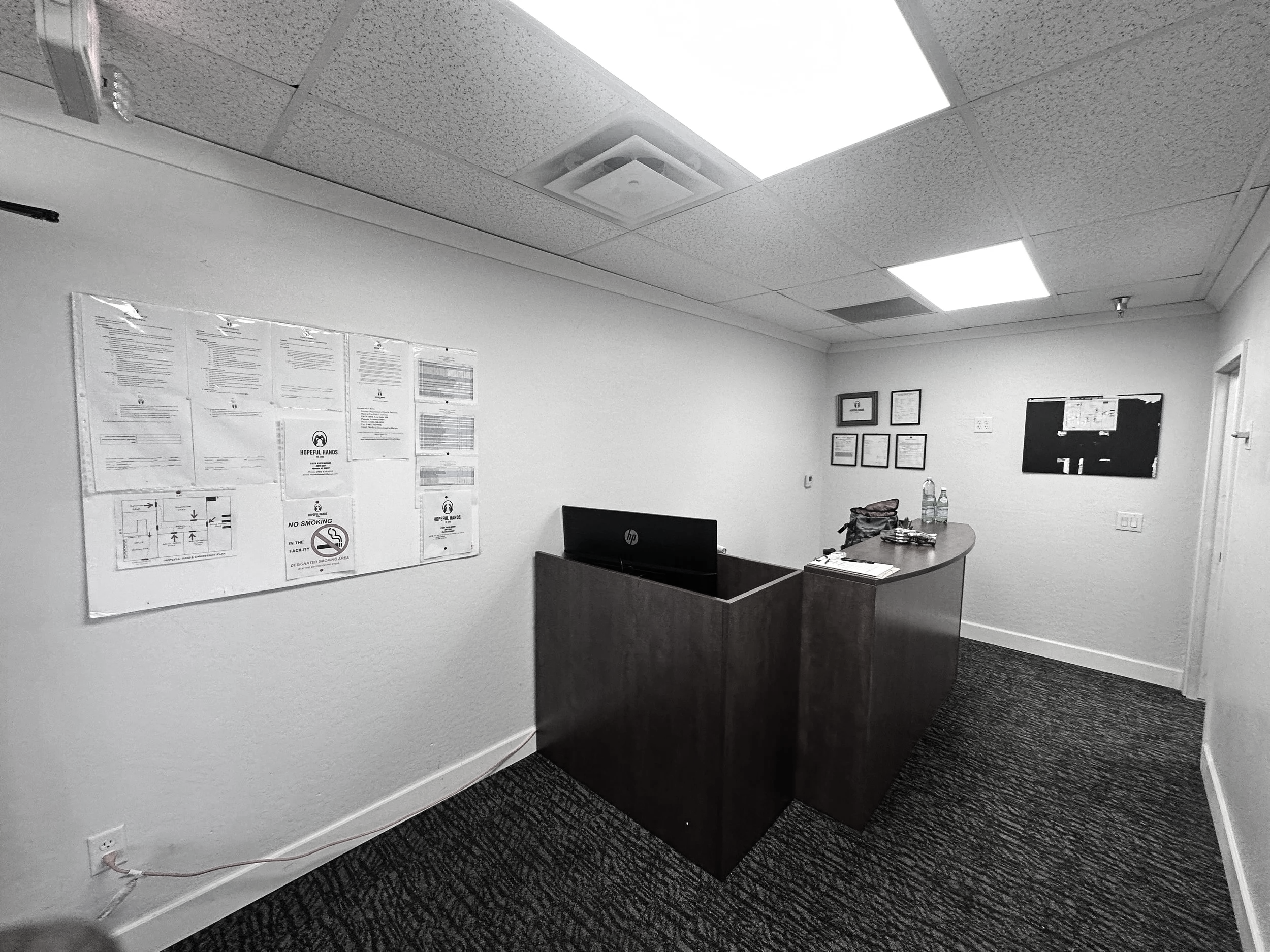 An office reception area with a wooden desk, a computer monitor, bottled water, papers, and framed certificates on the wall. There are ceiling lights and a white wall with various notices and a no smoking sign.