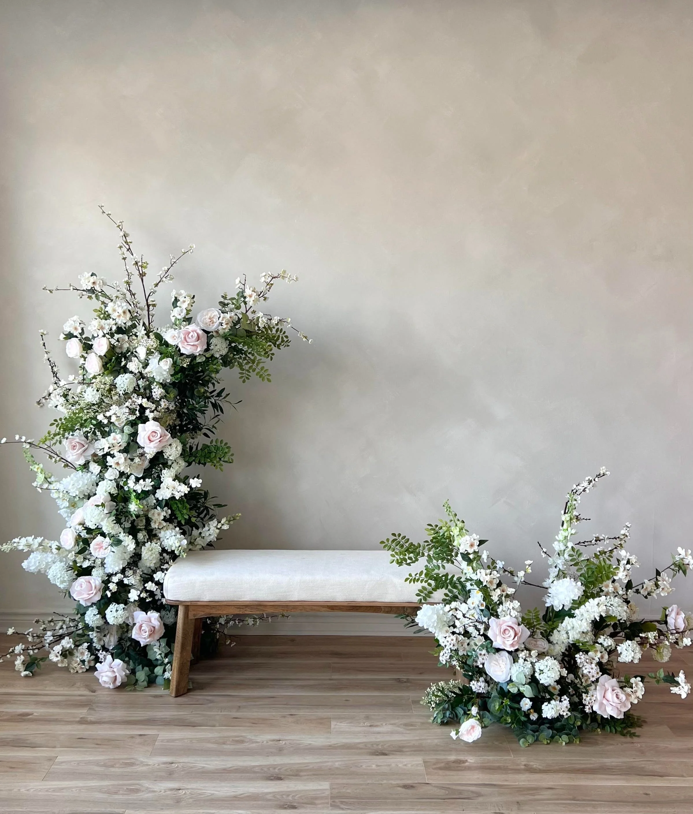 A white fireplace mantel with a gold-framed mirror on top, decorated with a white flower arrangement and four tall gold candlesticks with white candles, in a room with white walls and light wood flooring, reflected ceiling chandelier in the mirror.