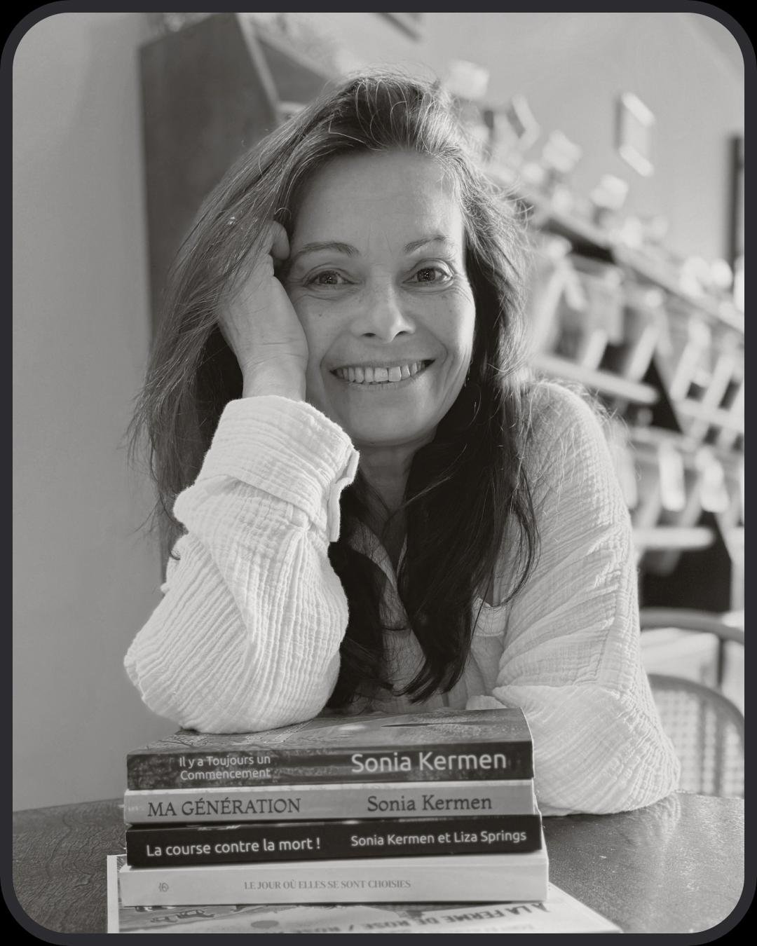 Photo en noir et blanc d'une femme souriante avec des livres devant elle, reposant sa tête sur sa main, dans un environnement intérieur.