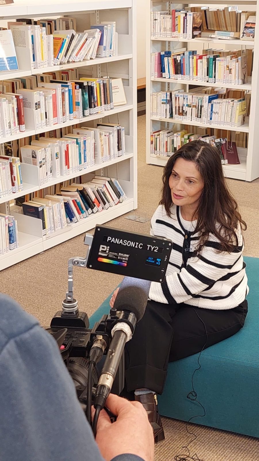 Une femme assise sur un sofa bleu dans une bibliothèque, en train d'être interviewée ou filmée par une caméra.