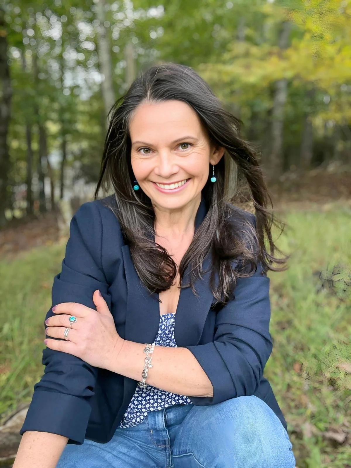 Une femme souriante avec des cheveux longs et noirs, portant une veste bleue et des bijoux, assise dans la nature, entourée d'arbres verts.