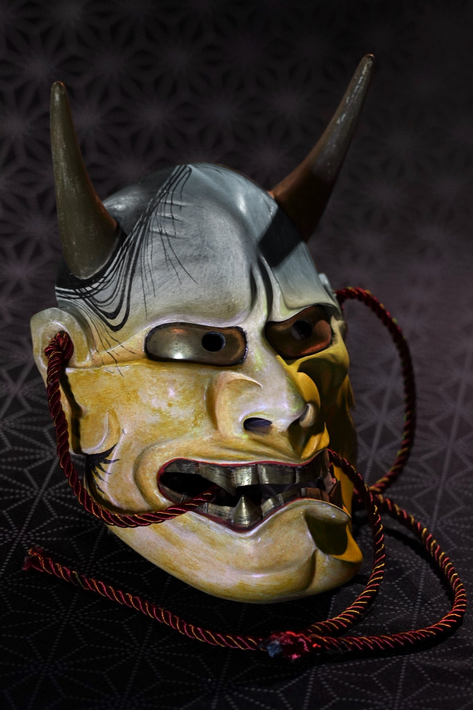 A traditional Japanese Hannya demon mask, featuring a fierce face with horns, a metallic mouth, and colorful cords on the sides, displayed against a textured dark background.