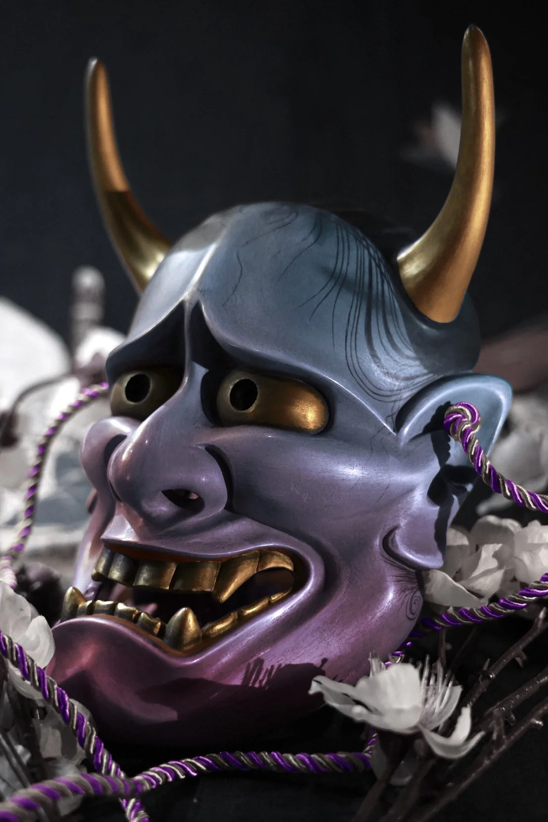 A detailed, colorful Japanese-style Hannya mask resembling a demon with horns, surrounded by white flowers and purple twisted cords.