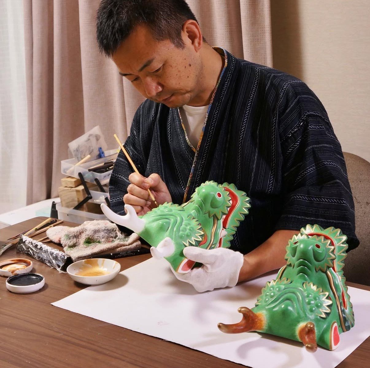 Hiromitsu painting a green dragon sculpture, seated at a table with painting supplies around him. The sculpture is part of a traditional decorative art piece.