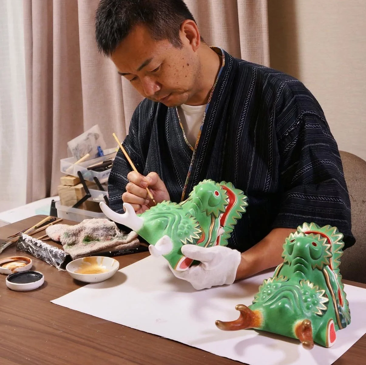 A man is painting a green dragon sculpture, seated at a table with painting supplies around him. The sculpture is part of a traditional decorative art piece.