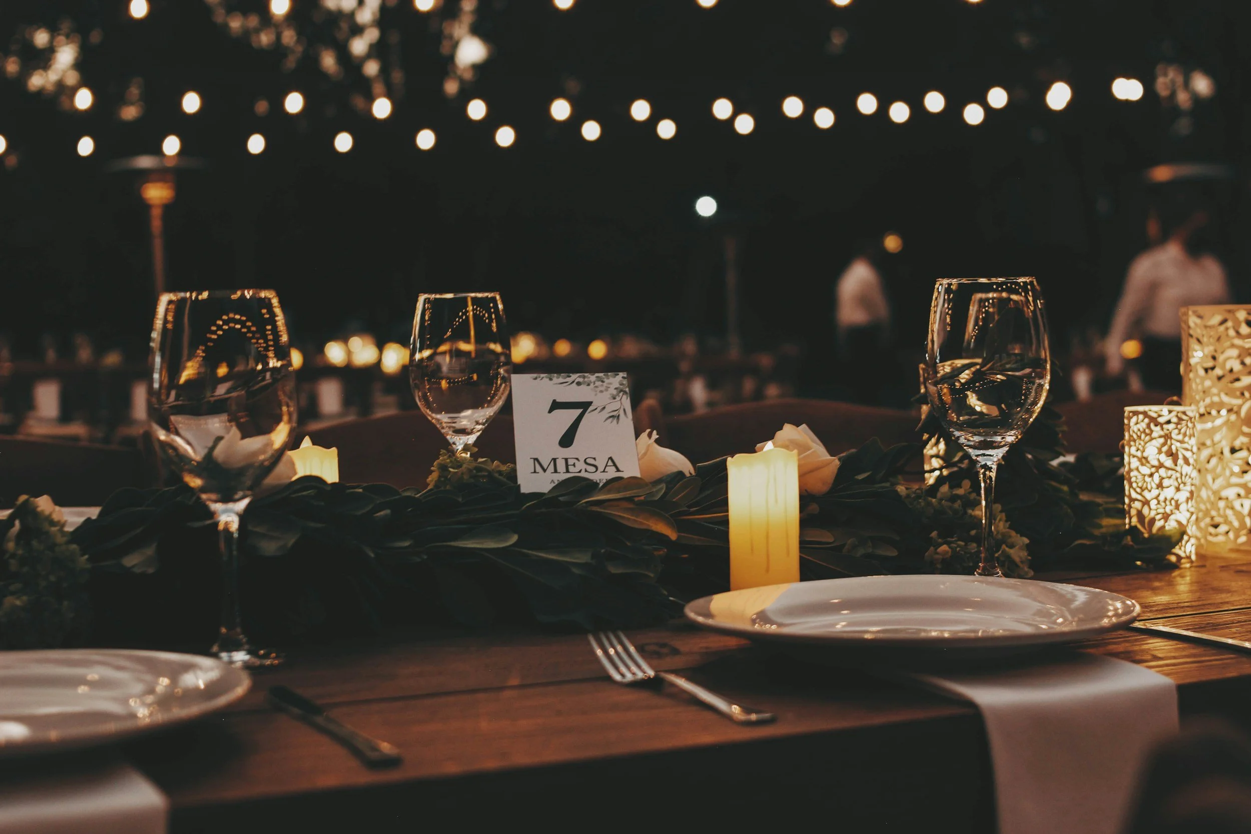 Wedding reception table decorated with candles, wine glasses, place settings, and a table number card that reads '7 MESA'. Blurred string lights are hanging overhead in a dark outdoor setting.