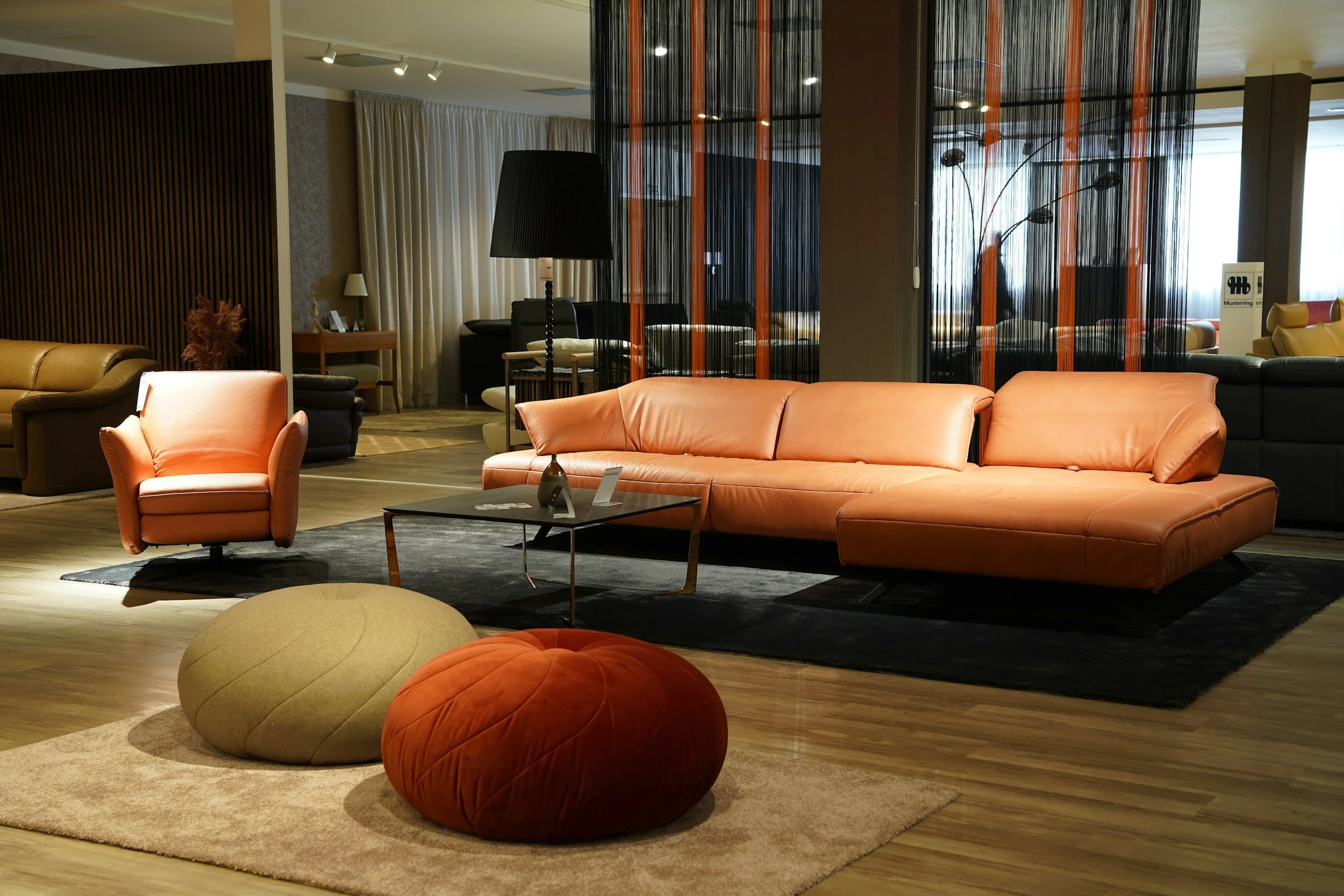 Modern hotel lobby with a pink sofa, orange and beige round ottomans, dark wood floors, and a black and orange decorative room divider. Armchairs, side tables, and lamps are visible in the background.
