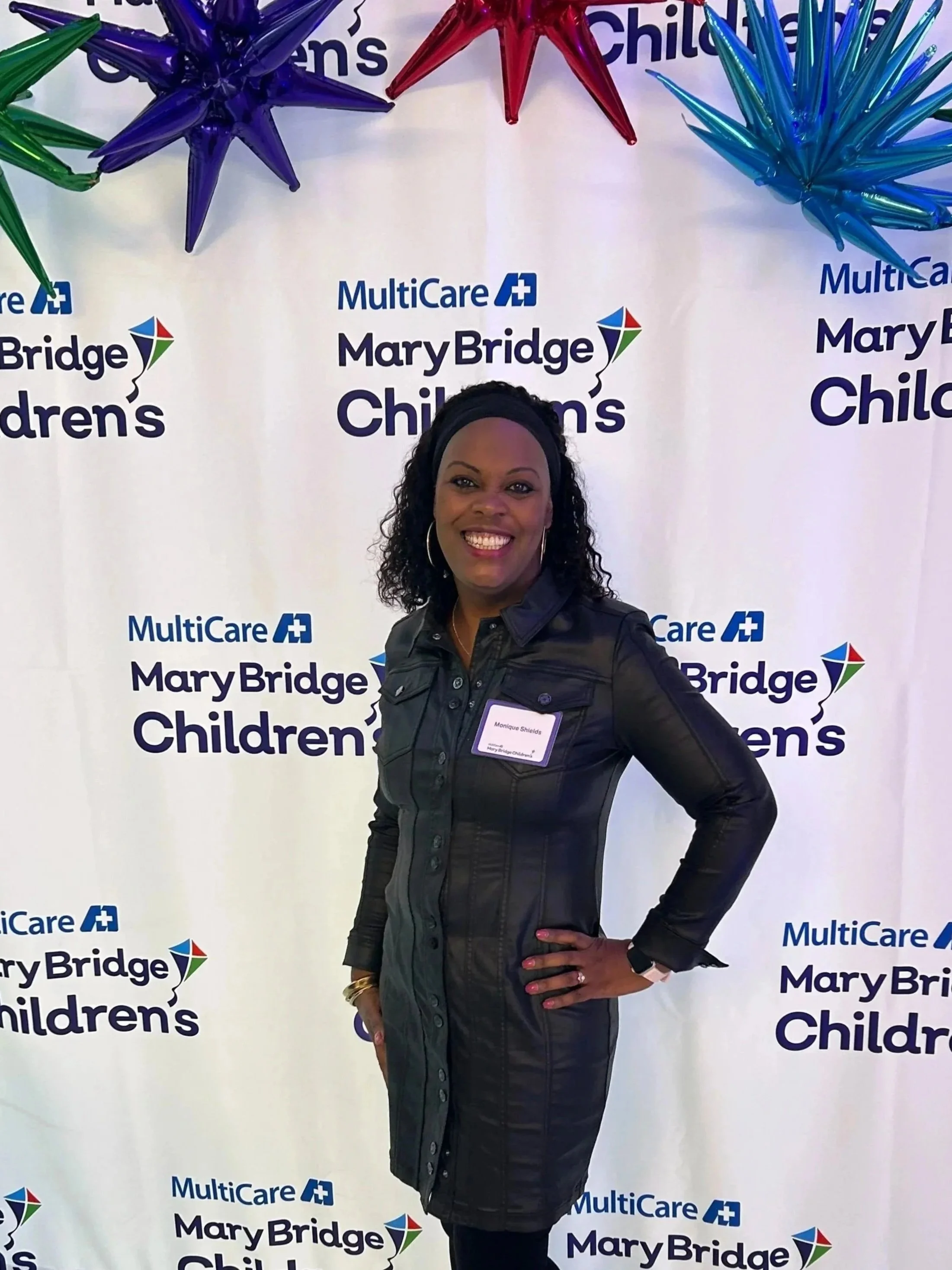Woman standing in front of a Mary Bridge Children's logo backdrop with colorful star decorations, smiling and wearing a dark jacket and earrings.