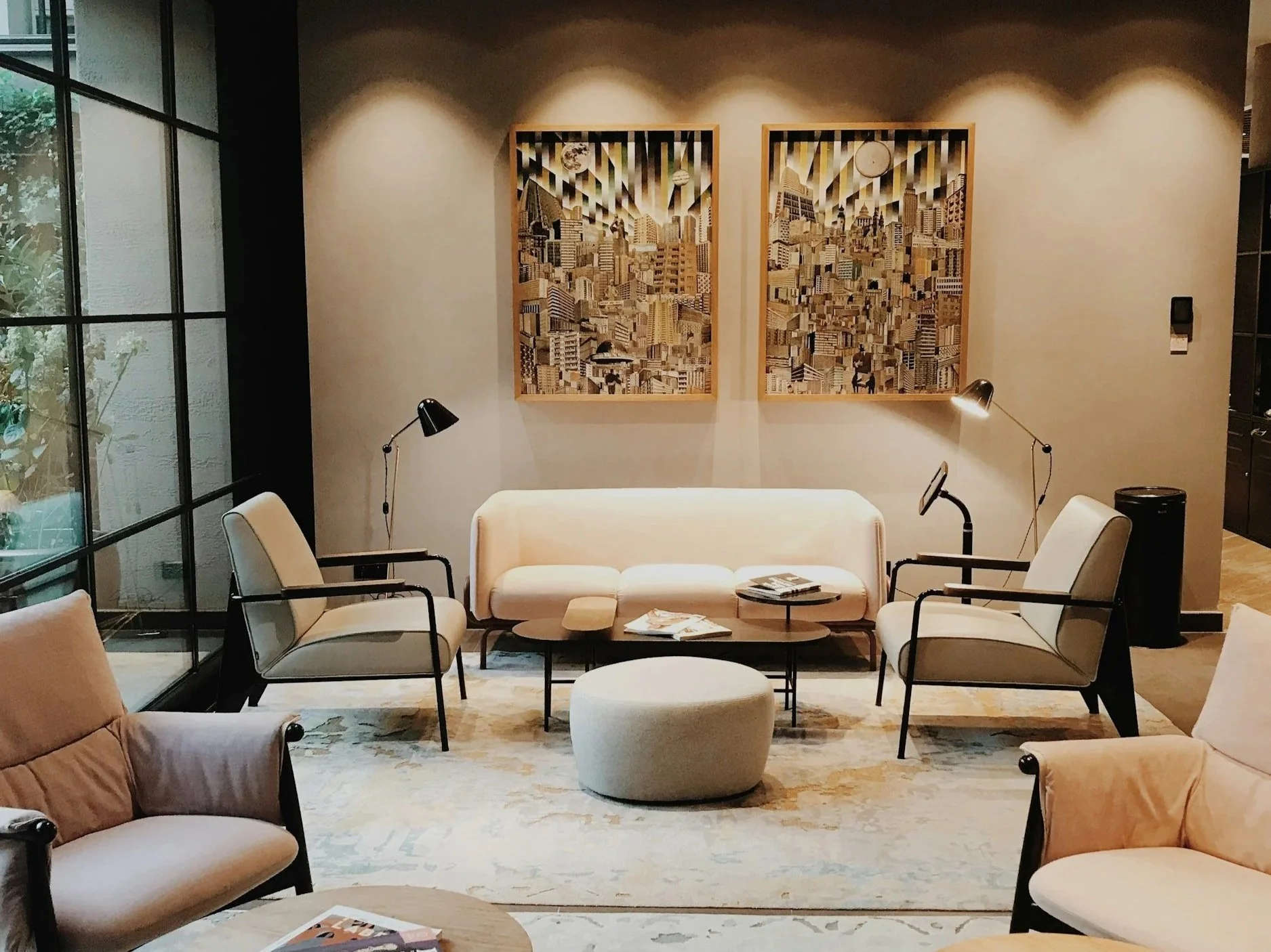 Modern hotel lobby with beige and cream seating, black metal frames, and two framed cityscape artworks on the wall. There are two black floor lamps and a round ottoman in the center, with natural light coming from large glass windows on the left.