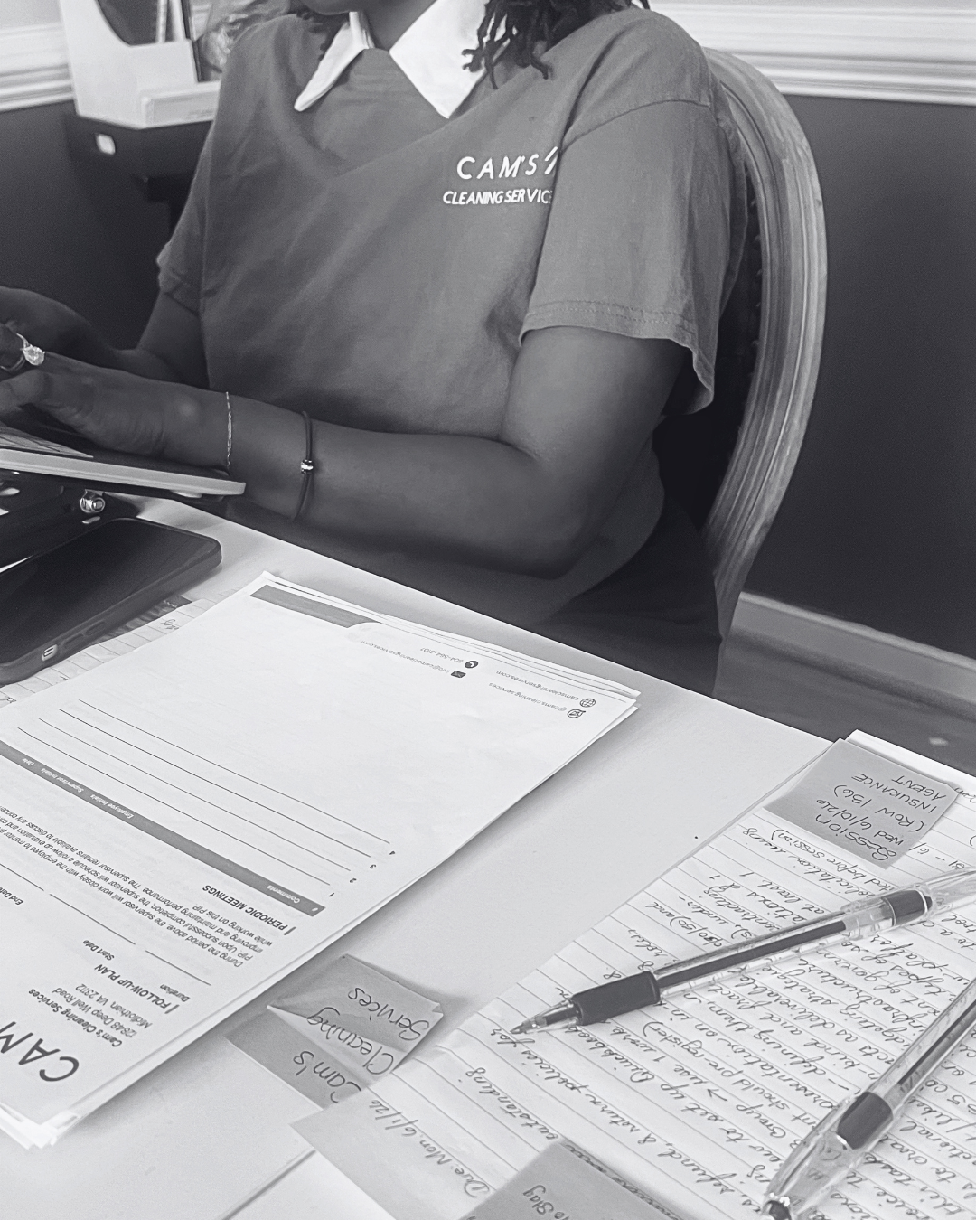 Conteh and Brown's HR Business Partner, in a Cam's Cleaning uniform, working at a laptop with a printed follow-up plan and handwritten coaching notes laid out on the desk.