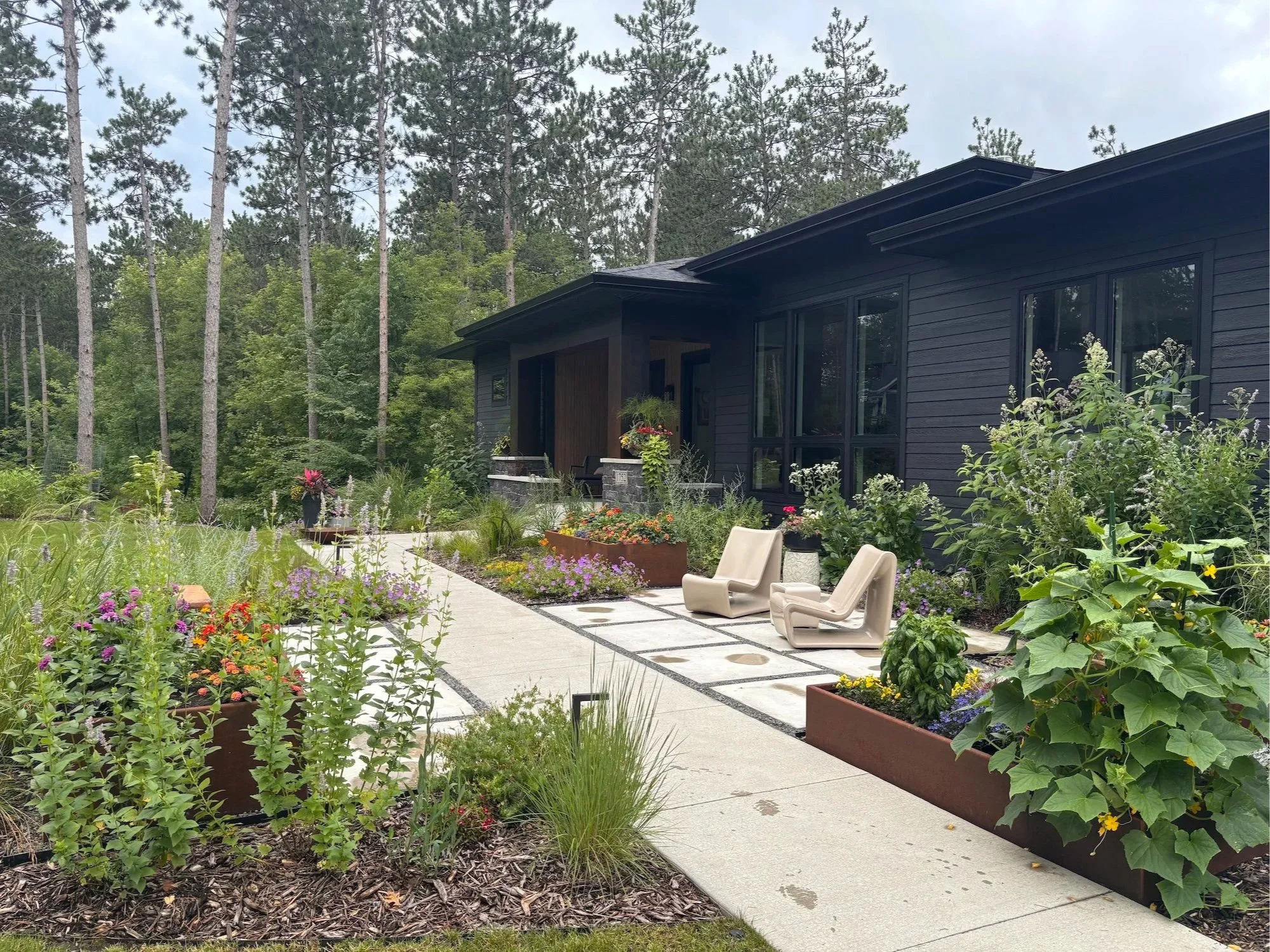 Modern seating patio set within native wildflower and grasses planting for pollinators, with steel raised beds for veggie and other edible plants such as herbs. This is an entrance landscape for a new home, which uses local materials for hardscape an