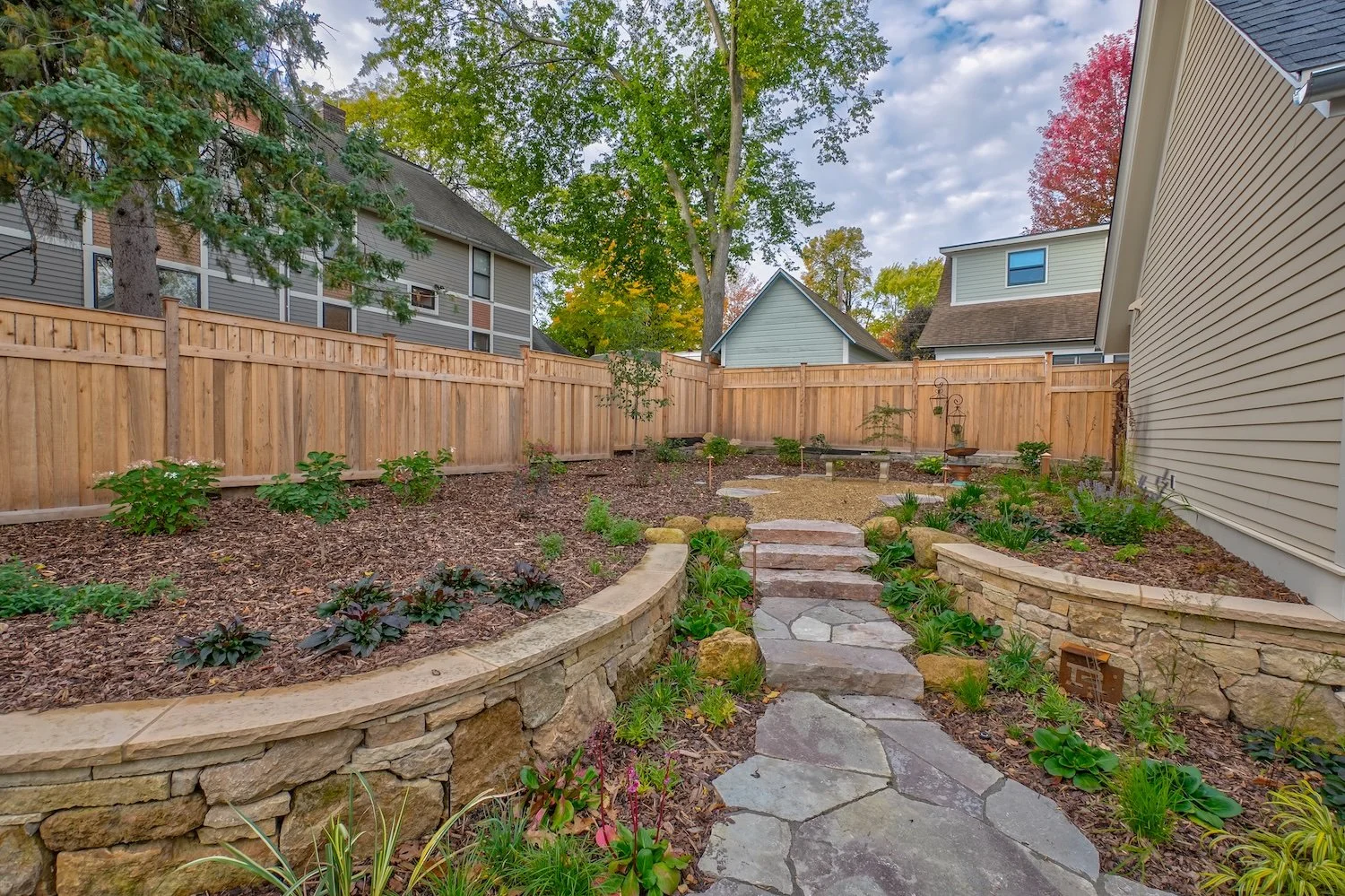 Professional residential landscape design showing a multi-level garden with limestone walls, flagstone steps, and newly planted shrubs in a heavily mulched bed under an autumn sky.