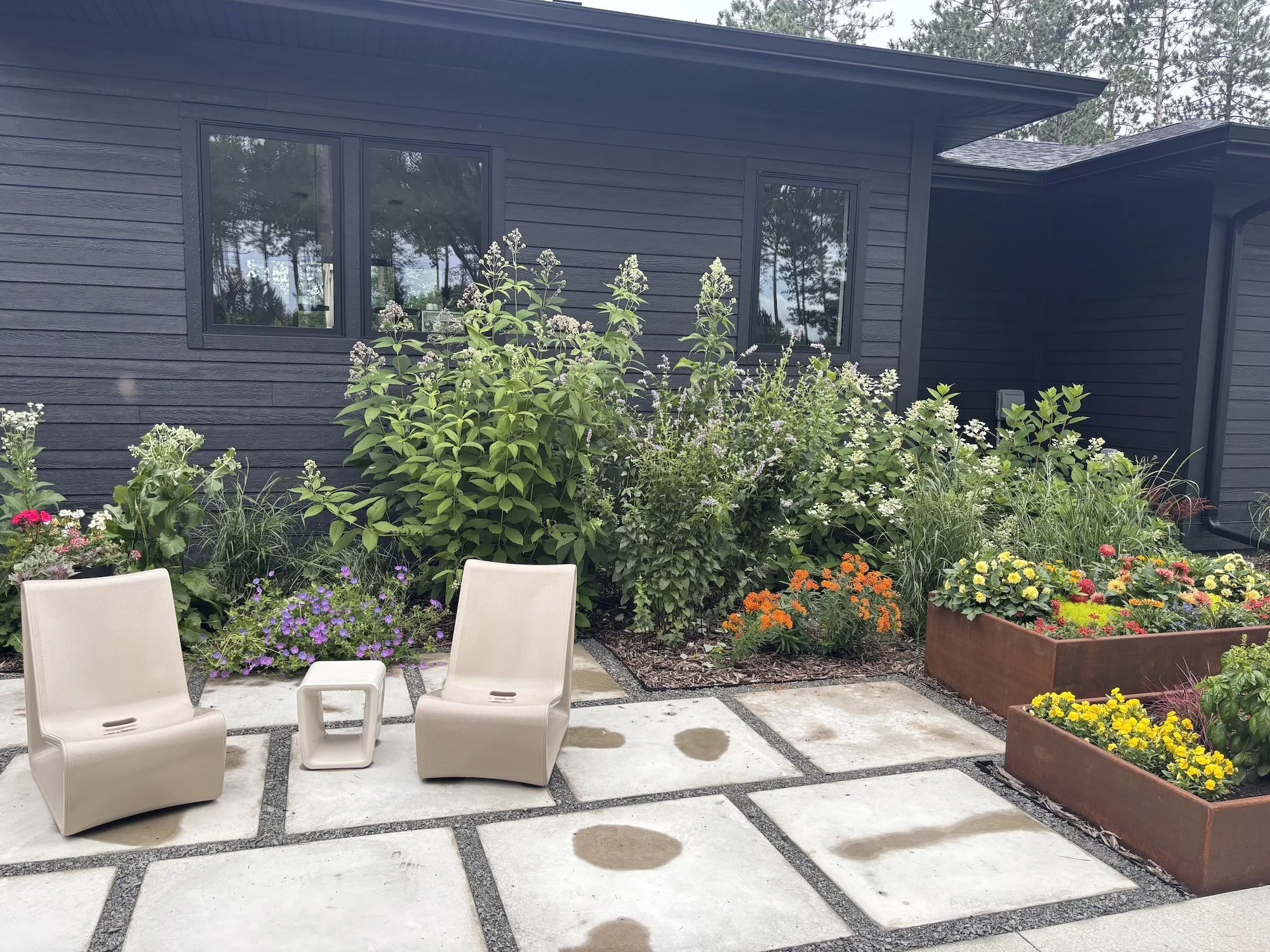 A closer shot of the modern seating patio, where the landscape homeowner sits with their morning coffee to listen to and watch songbirds, and see butterflies, bumblebees on the native wildflowers and colorful annual blooms. The locally made steel pla