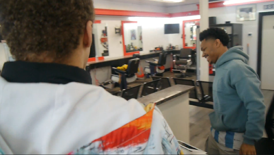 Two young men inside a barbershop, one in the foreground with curly hair wearing a white jacket with orange and yellow accents, and the other in the background in a blue hoodie and striped shorts, smiling and talking.