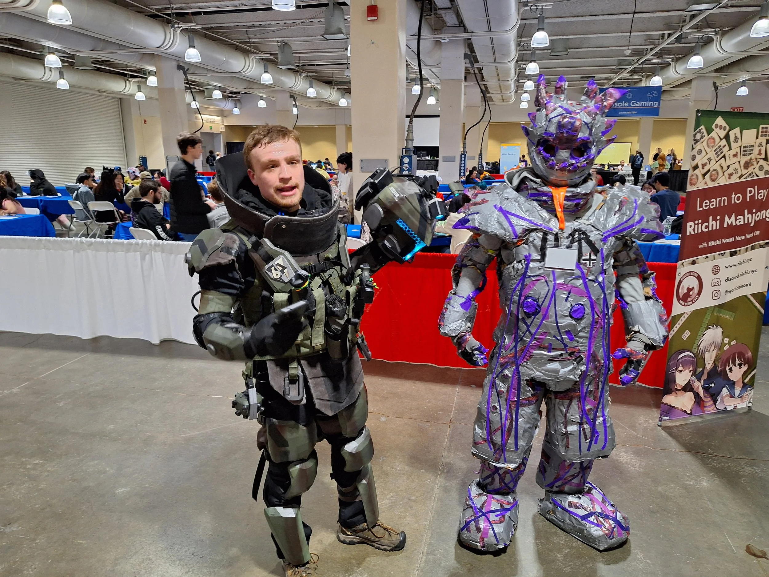 A man in a detailed, futuristic military-style costume next to a large robot-shaped costume made of aluminum foil and purple accents at a convention.