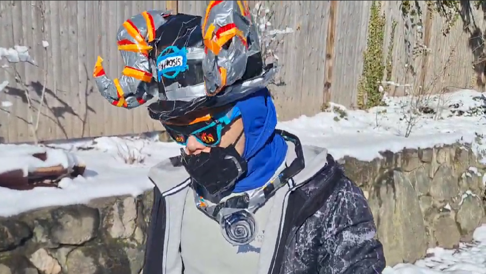 A person outdoors in winter gear, wearing a helmet decorated with a space-themed design, sunglasses, a face mask, a blue jacket, and gloves, standing in front of a snow-covered yard with a wooden fence and stone wall.