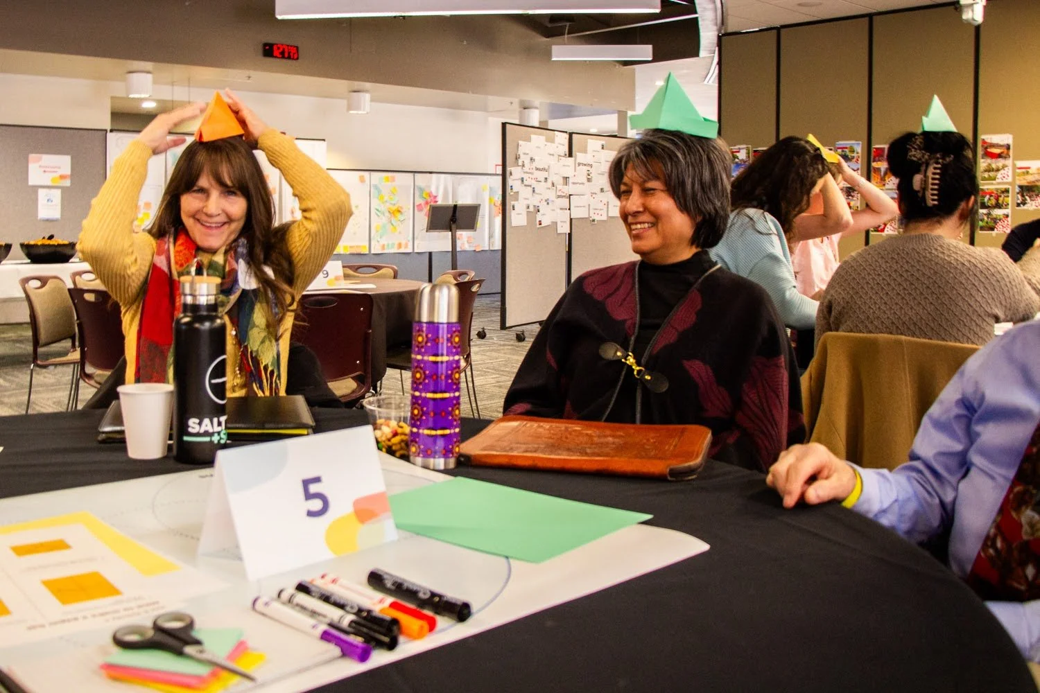 Participants at the Healthy & Thriving Places Summit sitting at a table with pens, markers, and construction paper, wearing colourful paper boats on their heads.