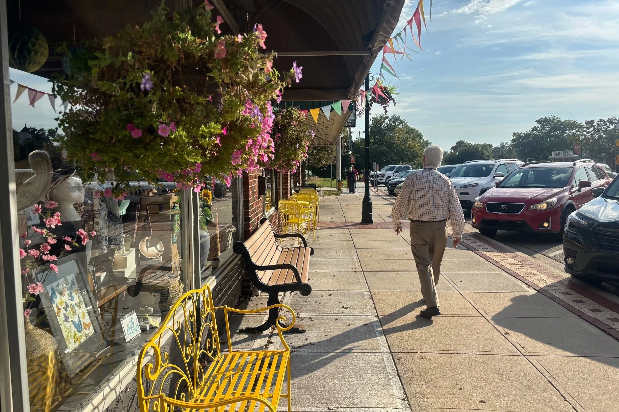 Photo of a wide sidewalk in overland park, with an older man walking down the street. The sidewalk is lined with small storefronts and benches, with decorative plants hanging from an awning above