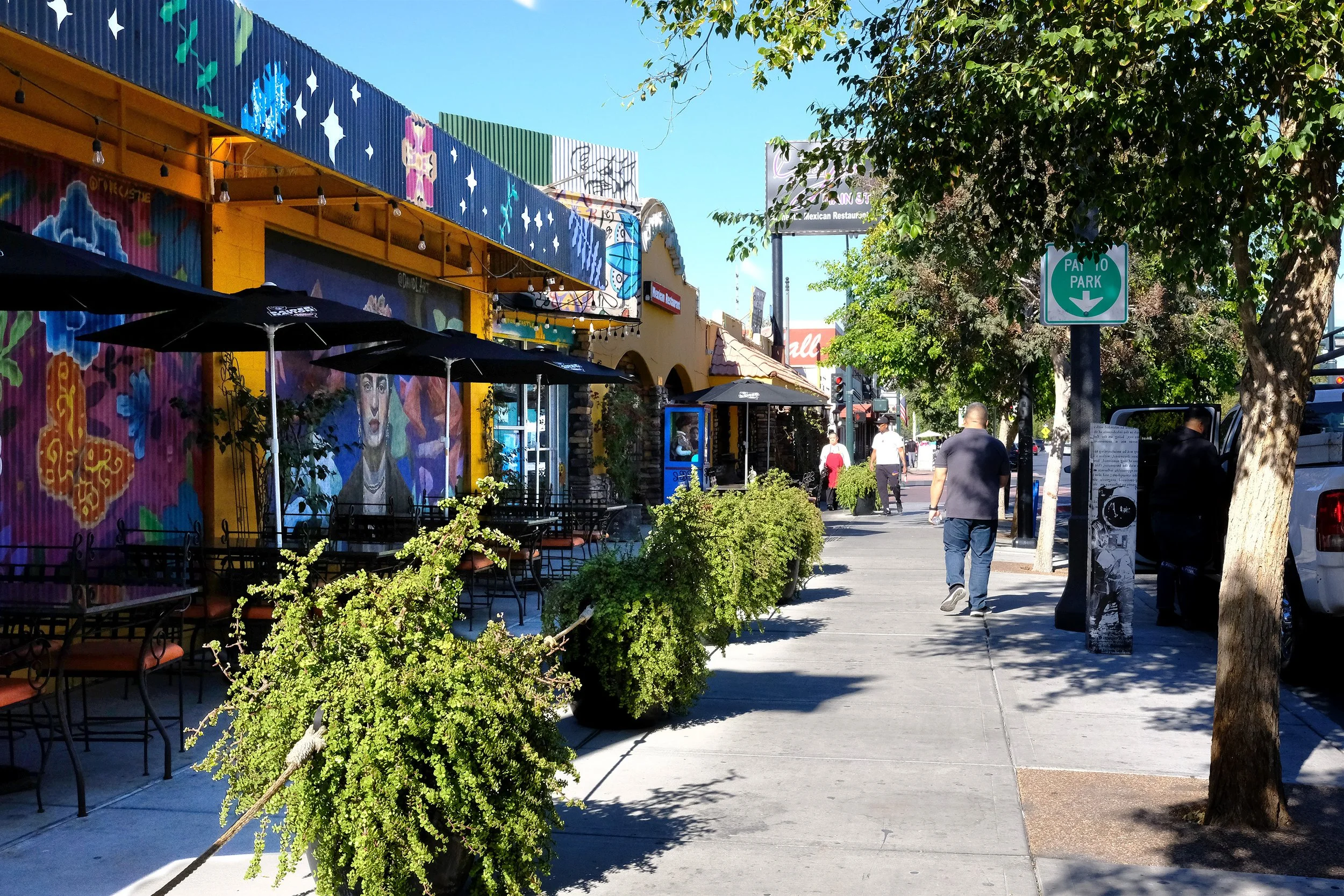 street-level photo of the arts district, with people walking along a wide sidewalk. The street is lined with restaurants and patios on one side, with large trees on the other side to offer shade and create a buffer between parked cars