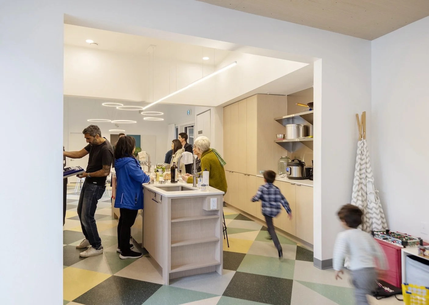 Photo of the common kitchen area at OUr Urban Village, a cohousing community. Residents of many ages gather round the kitchen island and socialize with neighbours