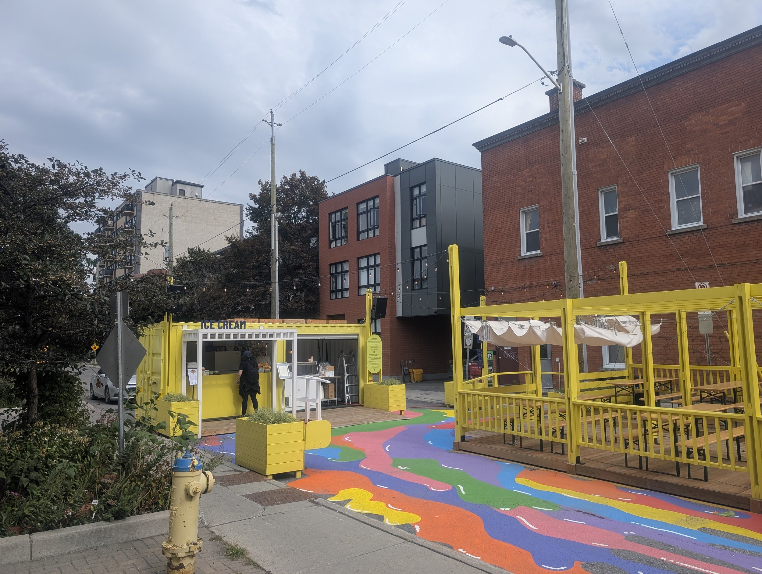 photo of the Frank St plaza, with a yellow painted shipping container selling ice cream, and a yellow fenced in patio area, and a colourful mural painted on the ground