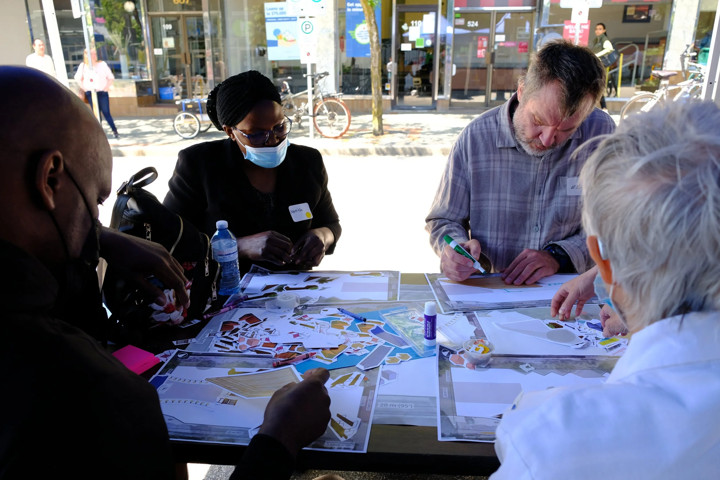 Photo of people sitting at an outdoor table during a workshop to co-design a plaza. There are cutouts of different seating and other public space elements on the table for participants to make a collage with