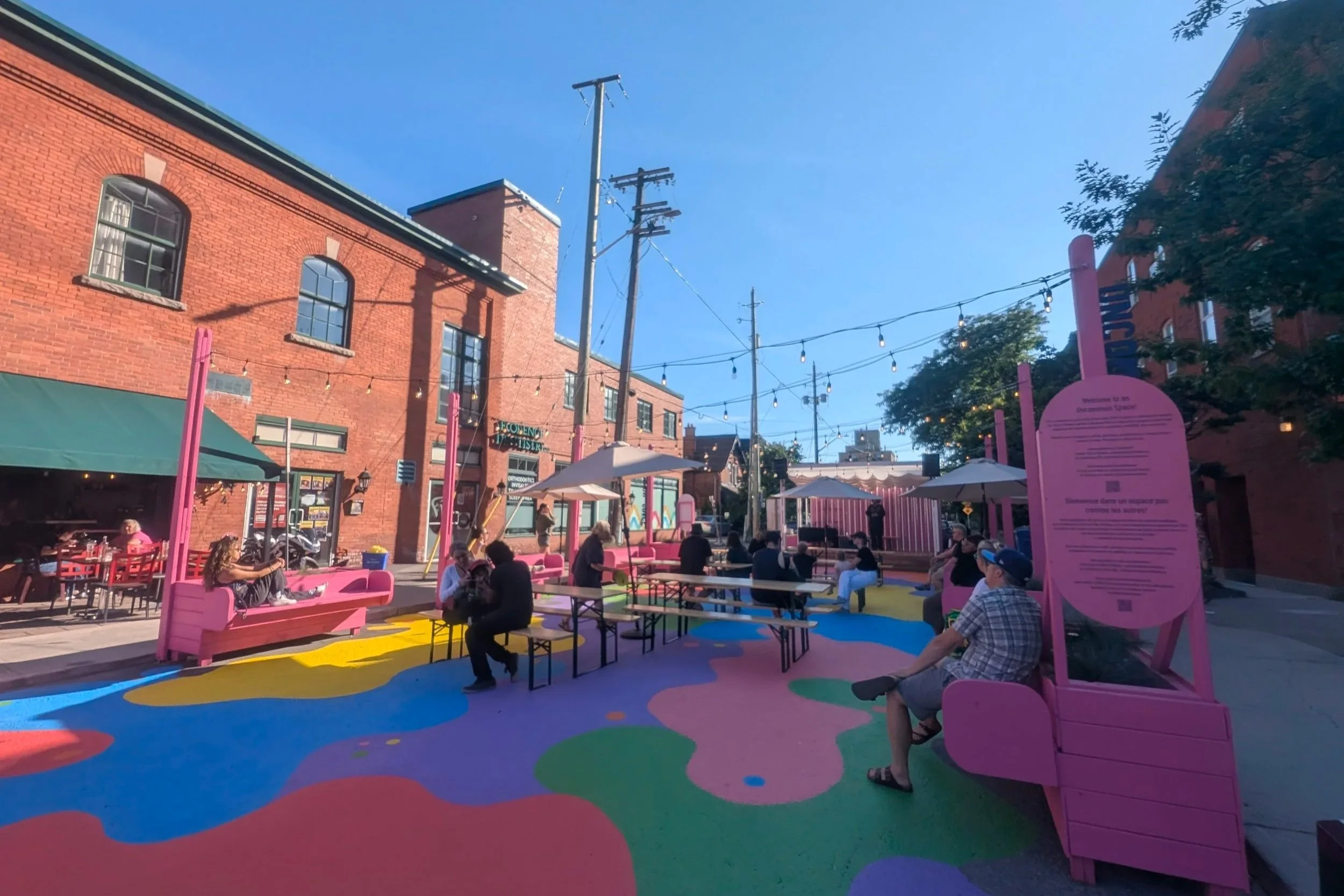 Photo of a street transformed into a plaza with a colourful mural painted on the ground, seating and planter boxes, umbrellas, and string lights overhead. people site in the plaza during a sunny day