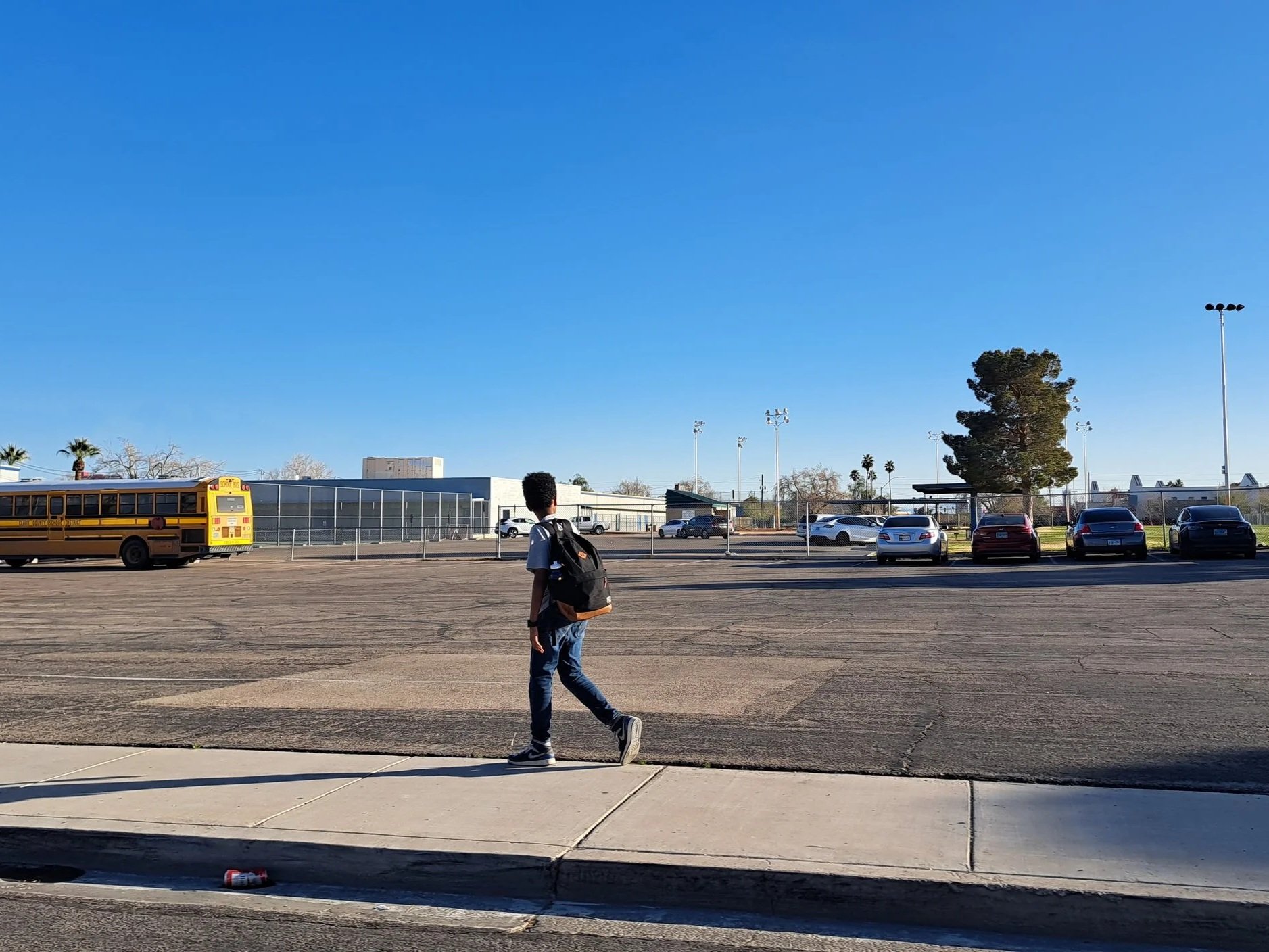 photo of a kid walking on a narrow sidewalk in the sun, beside a large open parking lot, with a school in the background