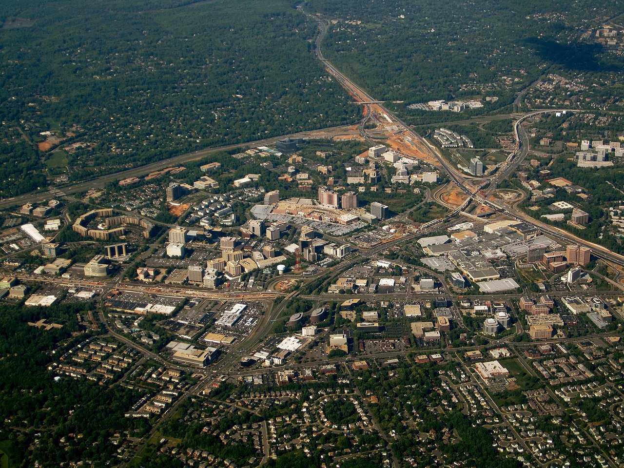 High-angle aerial view of Tysons Corner, Virginia, in 2010, showing a dense landscape of office buildings, shopping centers, and early construction of the Silver Line Metro. The area is surrounded by leafy, low-density residential areas